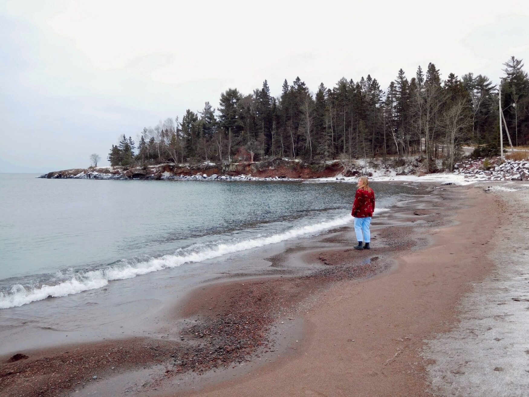 This image features a cheerful person standing on a sandy lakeside, partially covered with snow. They are dressed in a red patterned jacket, black top, light blue pants, and black boots. The sky is overcast, and the shoreline in the background has a mix of trees and rocky terrain, creating a peaceful and joyful vibe.