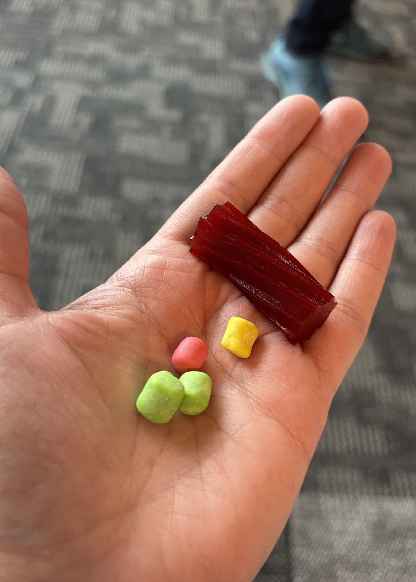 A close-up of a hand holding a selection of colorful candy pieces. The hand contains a small piece of red licorice and four coated candies in bright colors: green, pink, yellow, and another green one. The background shows a carpeted floor and part of a person's leg wearing dark pants and blue shoes.