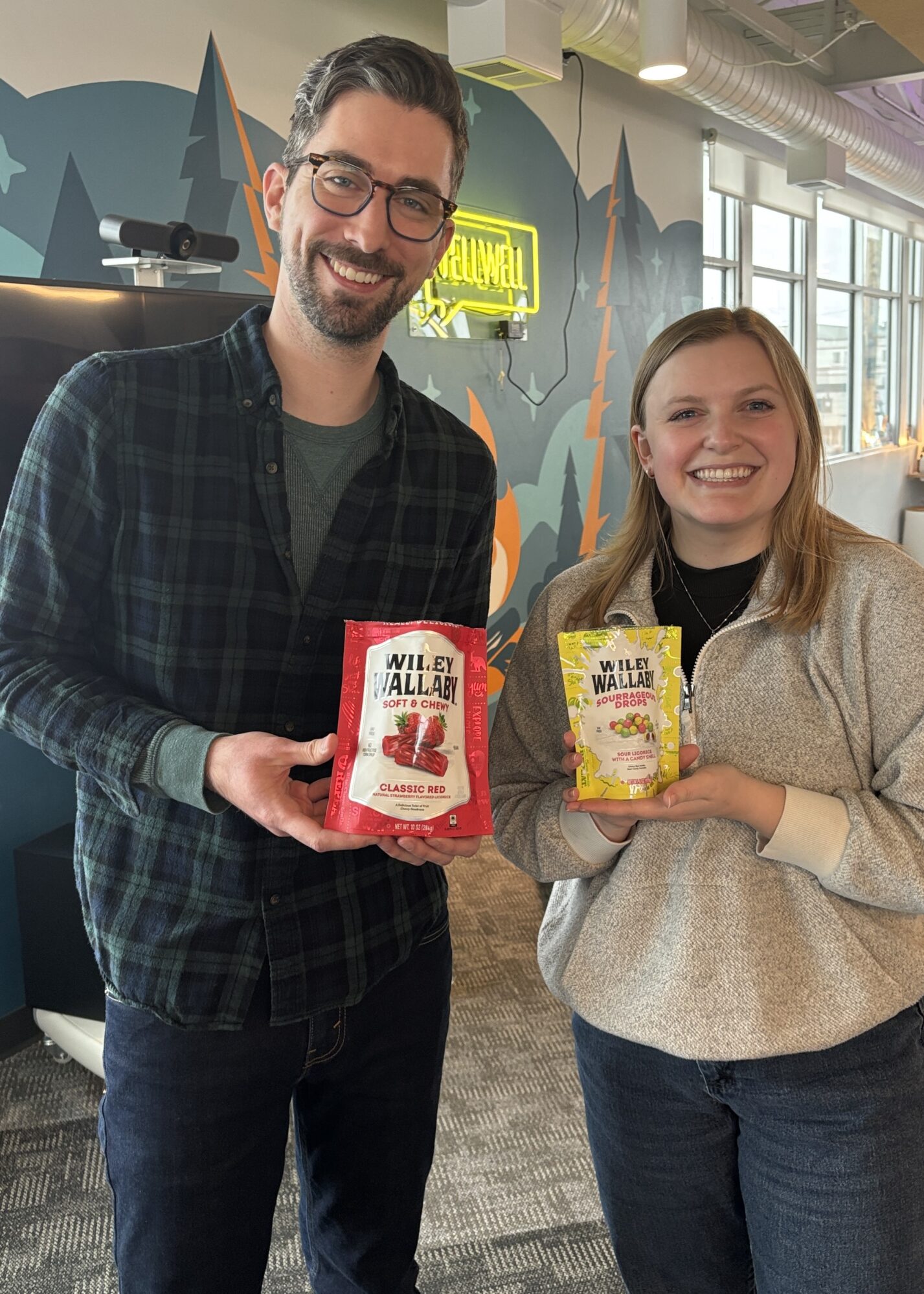 Two people standing indoors, smiling at the camera while holding colorful bags of Wiley Wallaby licorice. The person on the left is wearing glasses, a dark plaid shirt, and jeans, holding a red bag labeled "Classic Red." The person on the right is wearing a gray sweater and jeans, holding a yellow bag labeled "Sourrageous Drops." The background includes a brightly painted wall, a neon sign, and large windows letting in natural light.