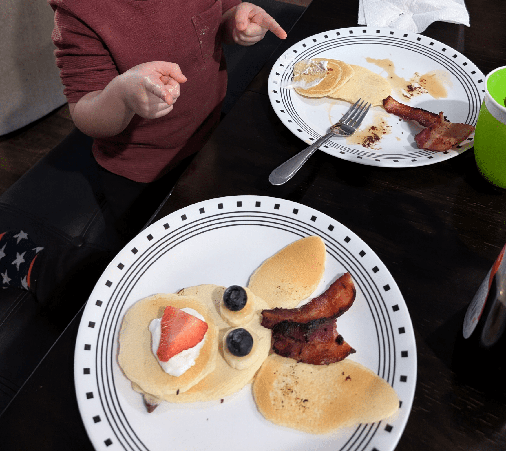 Two plates with pancakes, bacon, and fruit in the shape of a reindeer. There is a child pointing at both plates.