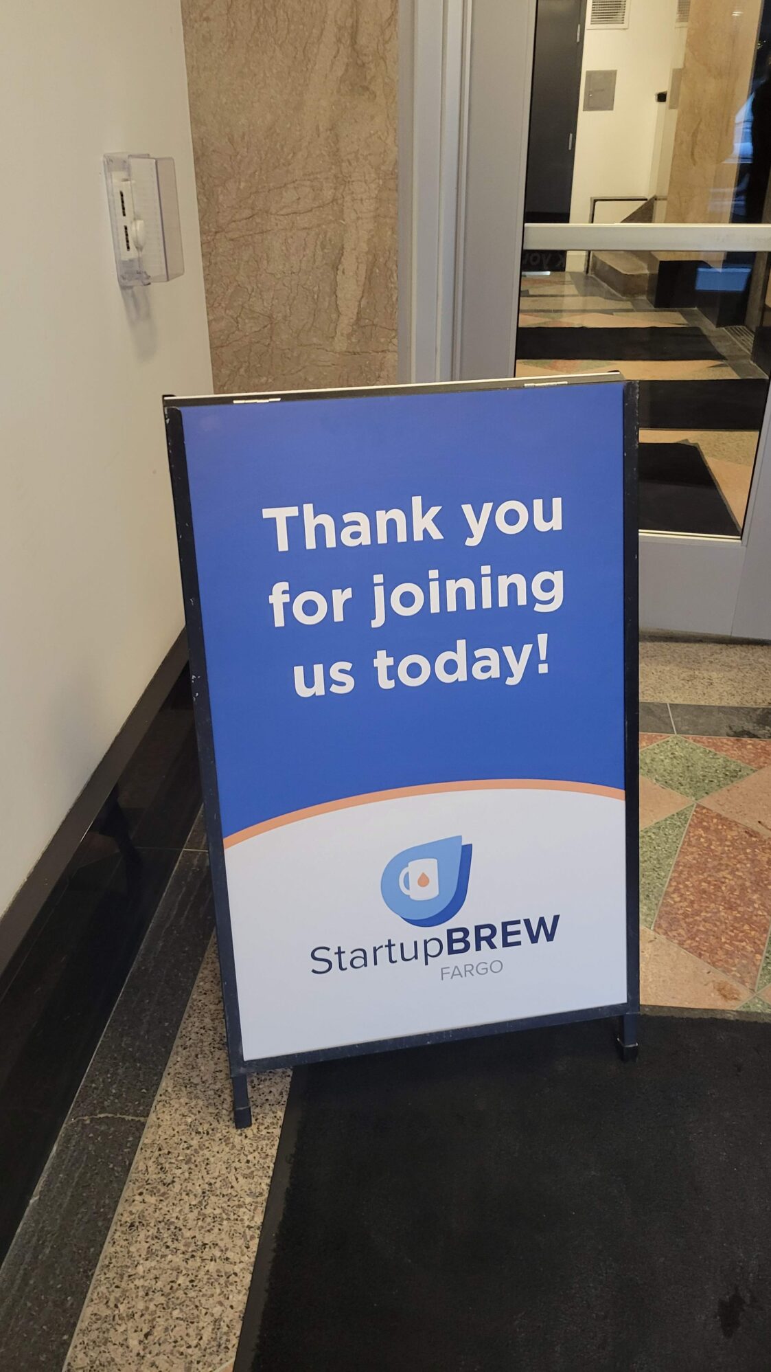 A blue and white sign reading "Thank you for joining us today!" with the StartupBREW Fargo logo is positioned in a hallway. The sign is freestanding on a black frame, and the background includes a marble wall and a glass door entrance.