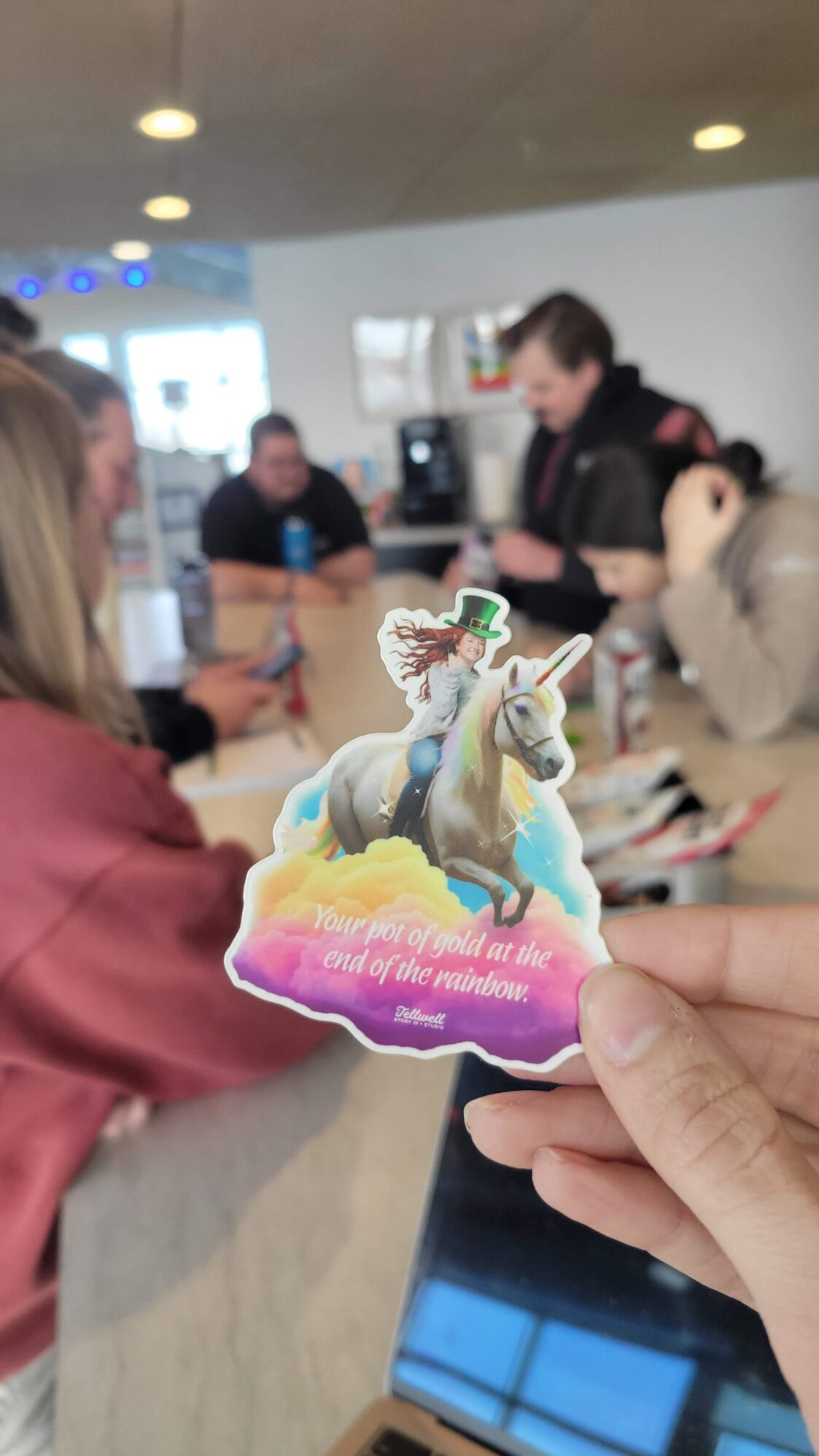 A person holds a colorful sticker featuring an illustration of a woman with red hair wearing a leprechaun hat, riding a unicorn through clouds. The sticker reads, "Your pot of gold at the end of the rainbow." In the background, a group of people is gathered around a table in a casual setting, engaging in conversation.