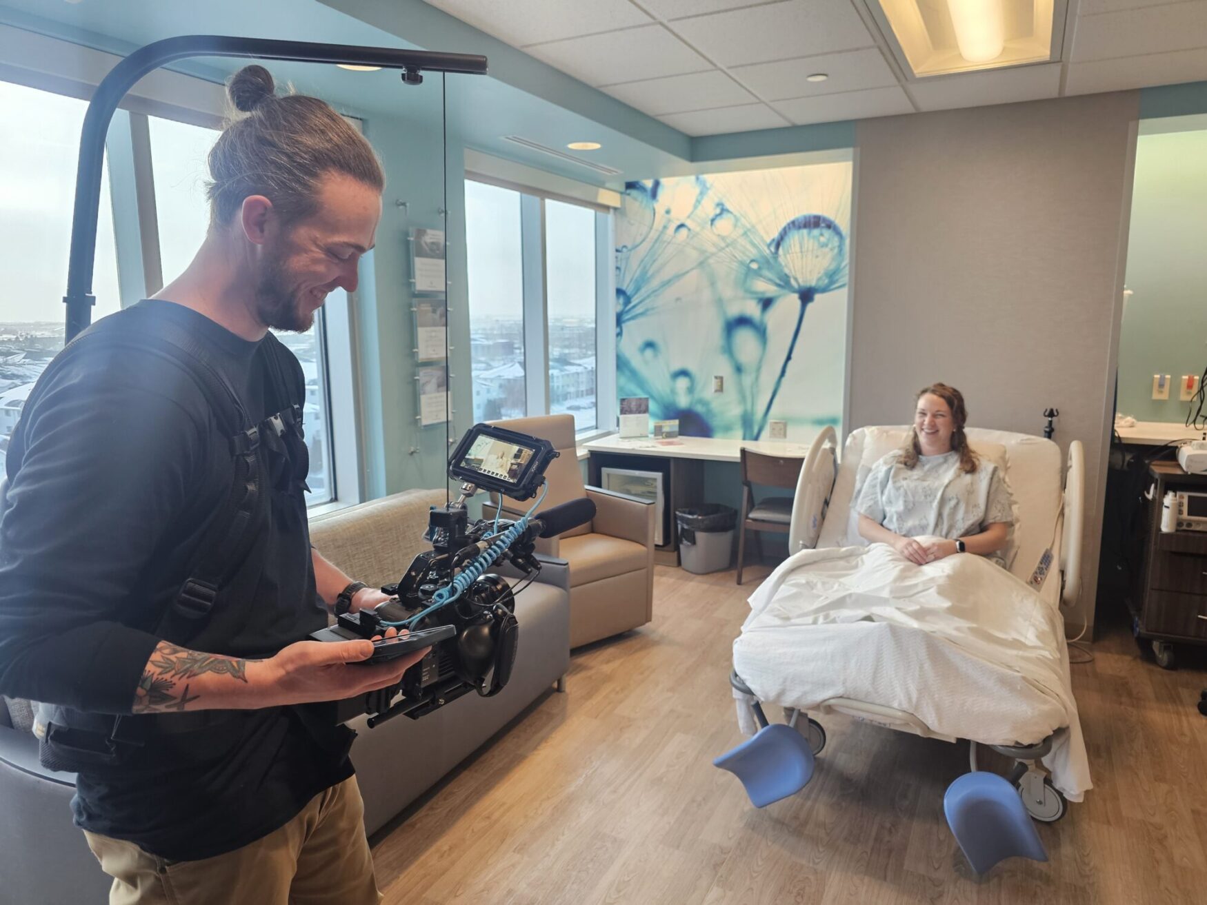 A videographer wearing a black shirt and a camera rig films a woman sitting in a hospital bed, wearing a hospital gown. The room is well-lit with large windows showing a snowy landscape, and a wall decoration featuring dandelion art is visible behind the patient.