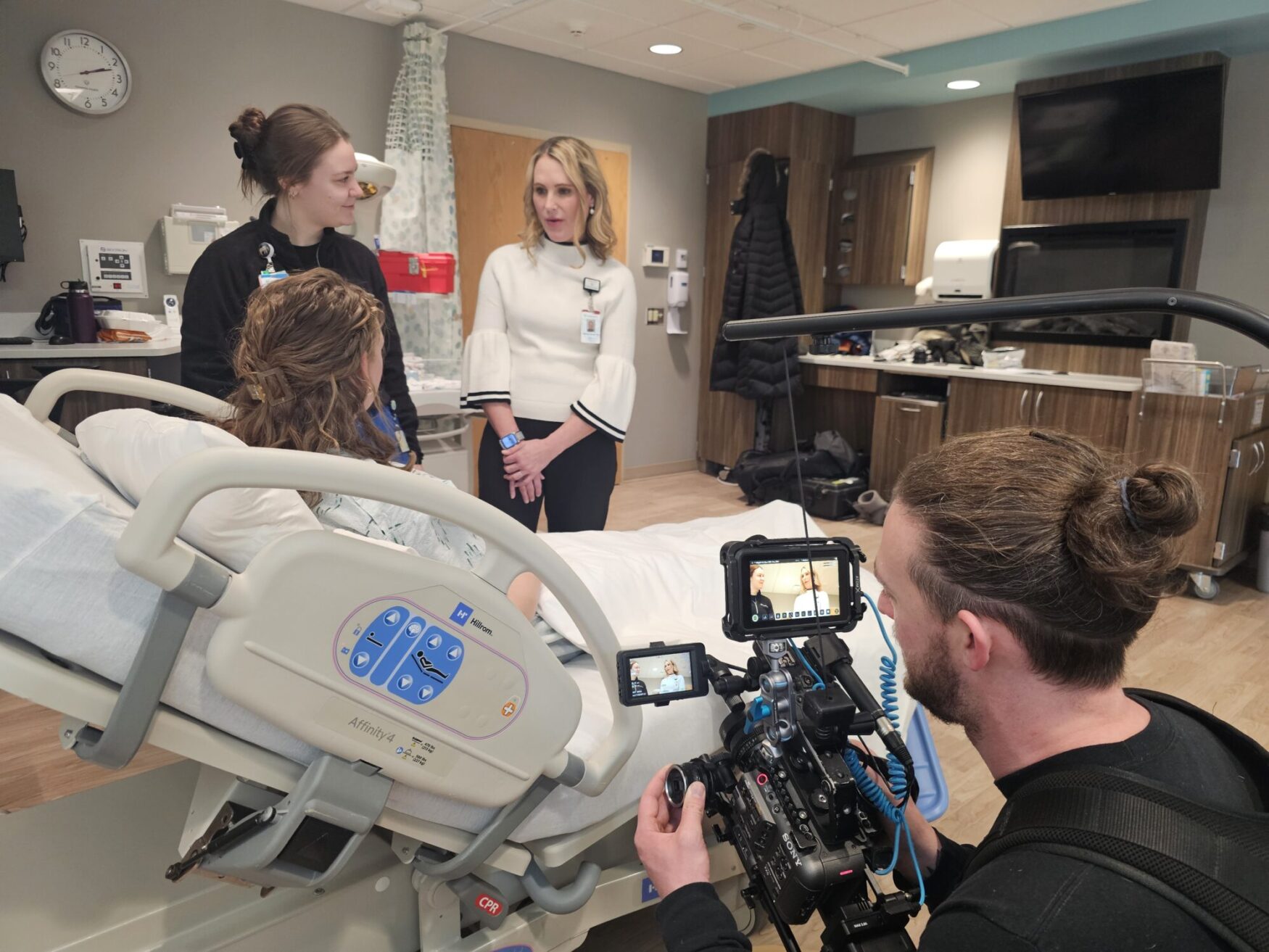 A hospital setting where a videographer captures a conversation between two medical professionals and a patient in a hospital bed. The videographer is using a camera rig with a monitor displaying the ongoing recording. The hospital room has wooden cabinets, medical equipment, and a clean, professional appearance.