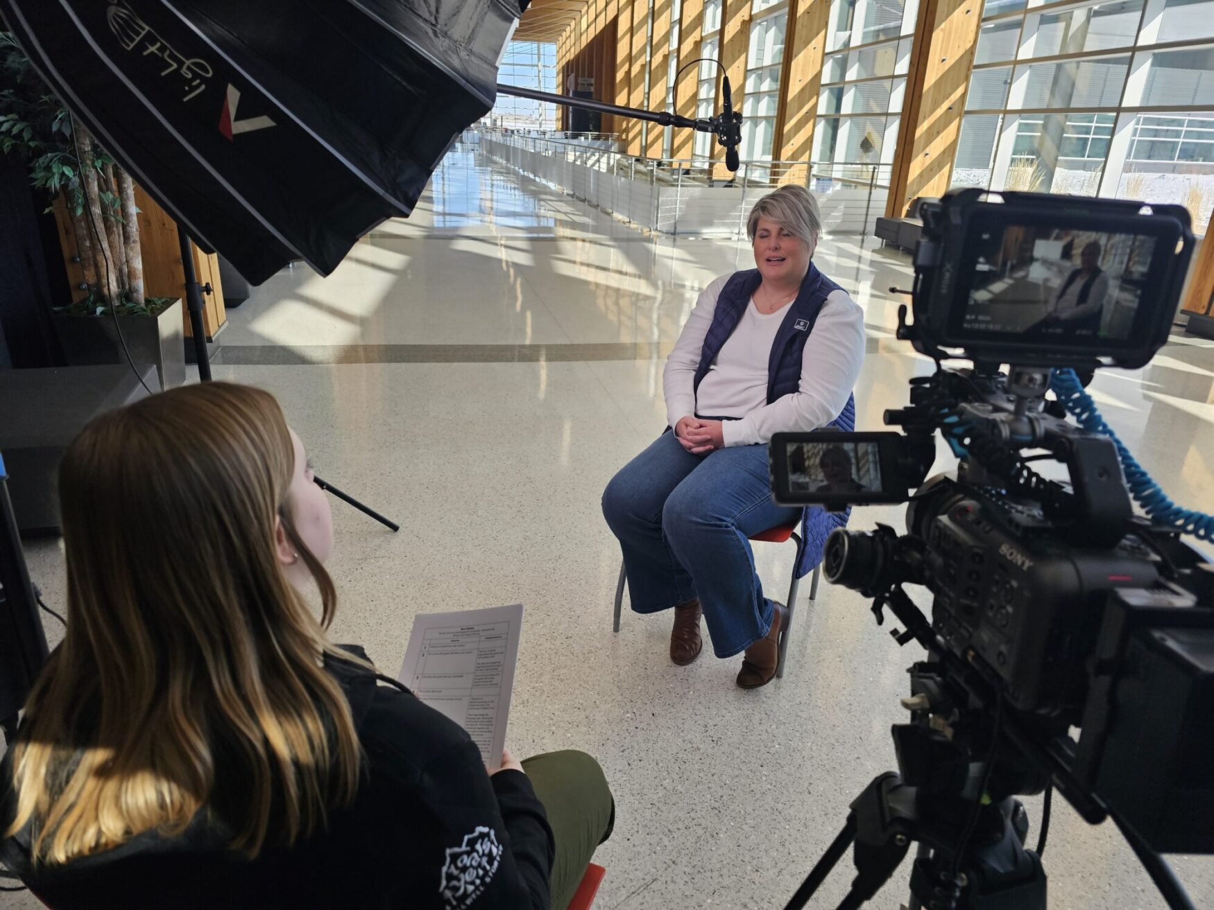 This image captures a professional interview setup in a modern, well-lit indoor space with large windows. A woman in a white shirt and blue vest sits on a chair while being interviewed. The interviewer, seen from behind, holds a sheet of paper with questions. A professional video camera is recording the scene, with a small monitor displaying the interviewee. Additional equipment, including a boom microphone and lighting, is set up for optimal audio and video quality.