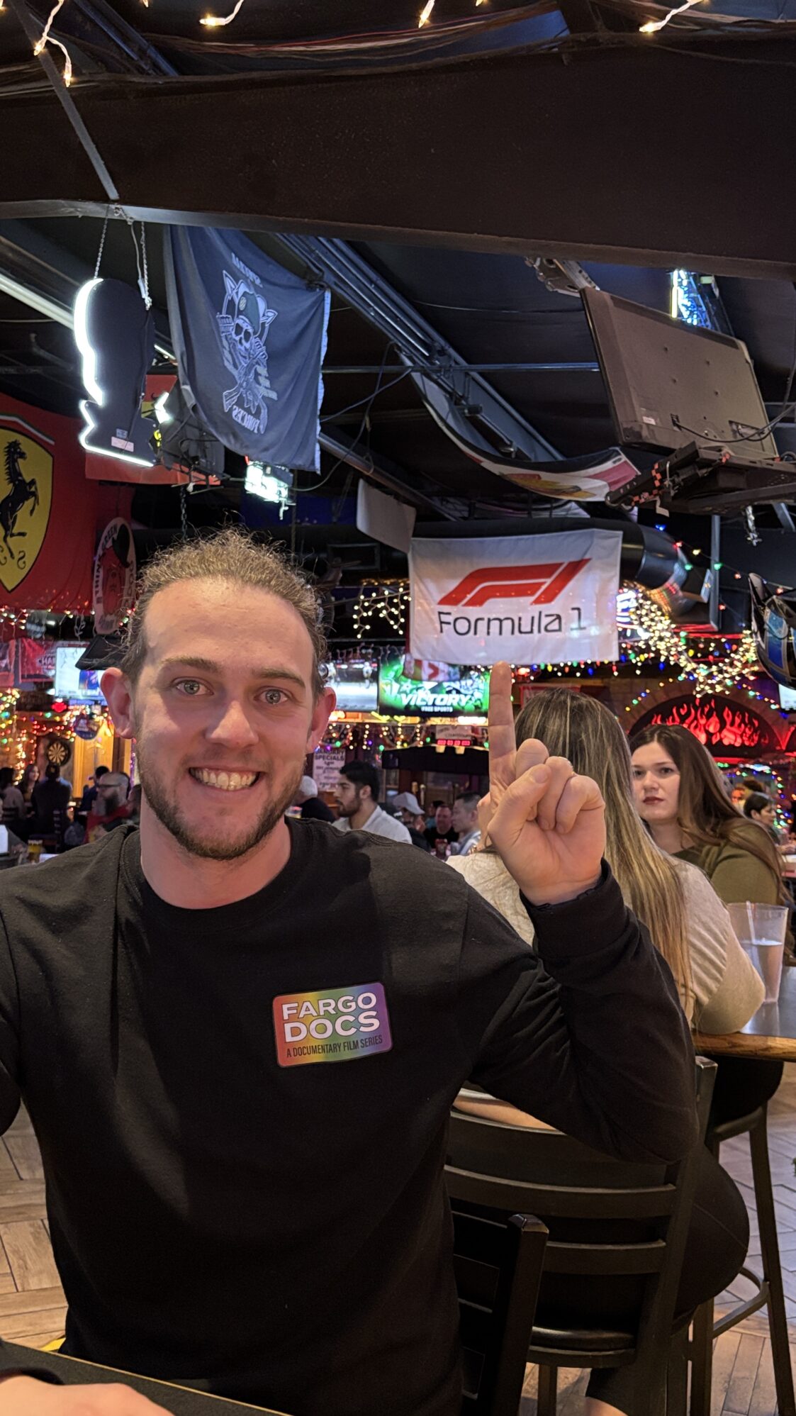 A smiling man with light skin, hair pulled back in a bun, and a beard is sitting at a bar or restaurant, pointing up at a Formula 1 flag hanging from the ceiling. He is wearing a black long-sleeve shirt with a colorful "Fargo Docs: A Documentary Film Series" logo on the chest. The background is filled with festive string lights, multiple TV screens, and various flags and decorations, including a Ferrari logo banner and a pirate flag. Other patrons are visible in the background, engaged in conversation and dining.