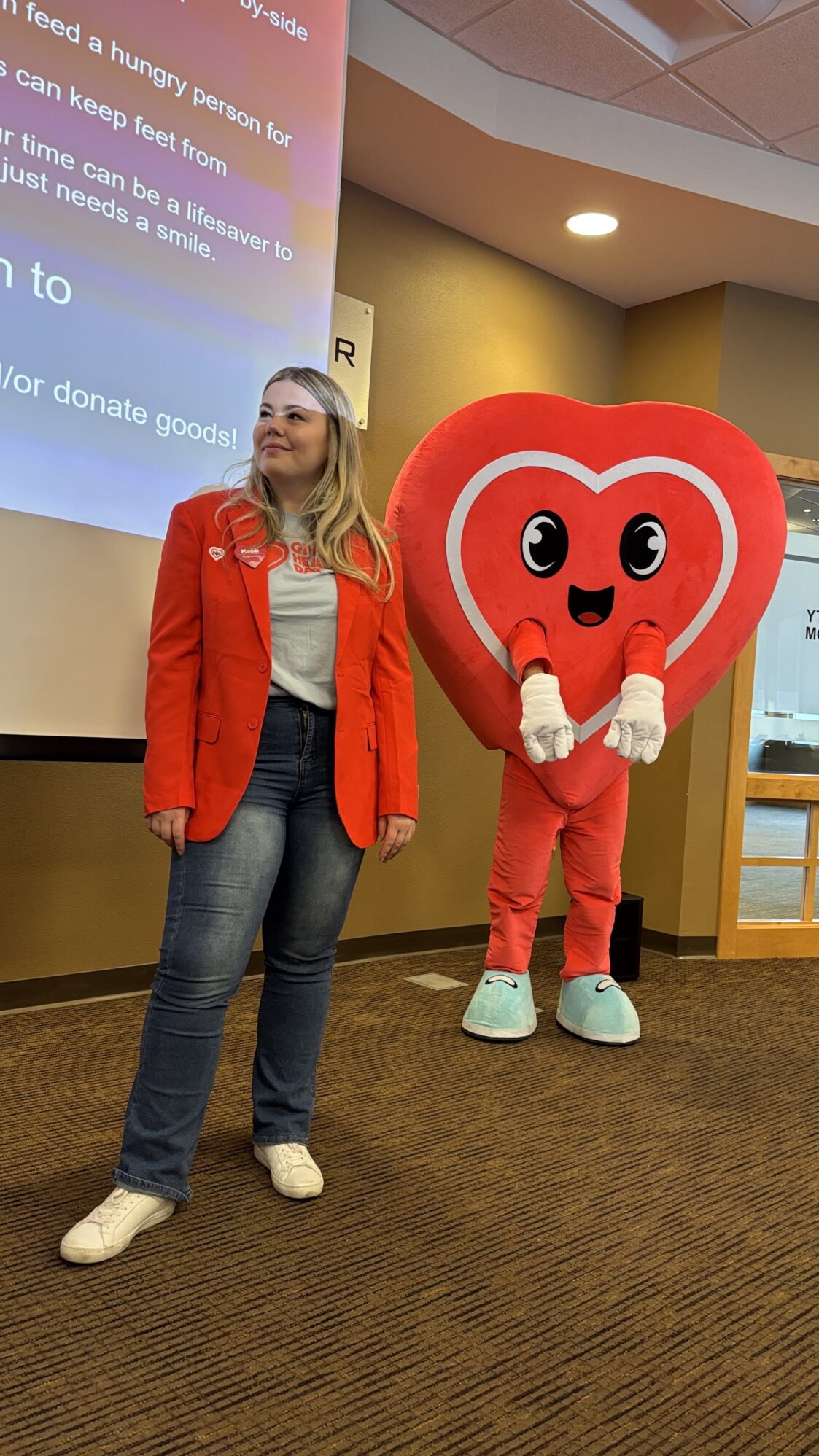 A woman in a red blazer and white t-shirt stands in front of a large presentation screen with a projected slide, speaking to an audience. Beside her is a mascot dressed in a red heart costume with big eyes, a smiling face, and white gloves. The room has warm lighting.