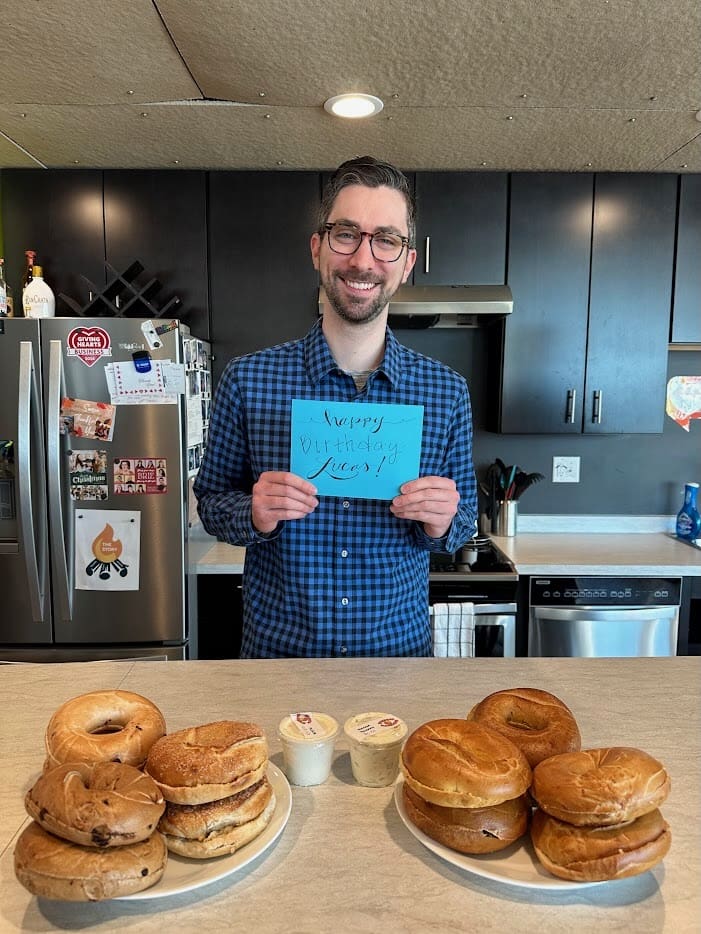A man with short brown hair, glasses, and a beard stands in a kitchen, smiling at the camera. He is wearing a blue plaid shirt and holding a handwritten sign on blue paper that says "Happy Birthday Lucas!" In front of him on a light-colored kitchen counter are two plates filled with assorted bagels and small containers of cream cheese. The background features a modern kitchen with dark cabinets, a stainless steel refrigerator covered in magnets and photos, a stove, and various kitchen utensils.