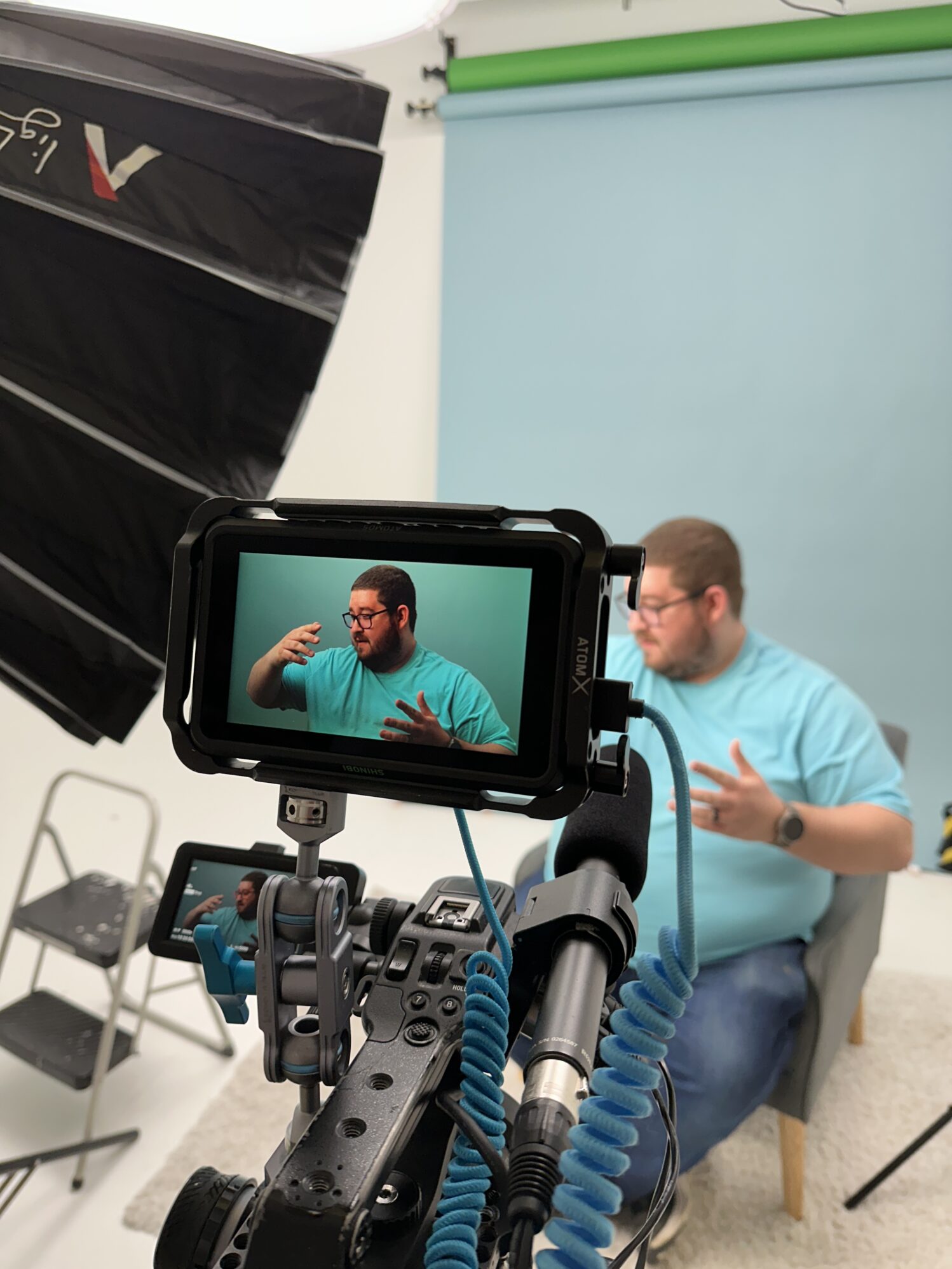 A behind-the-scenes look at a video production setup in a studio. A man wearing a light blue t-shirt sits in front of a light blue backdrop, gesturing as he speaks. The camera in the foreground captures him on a small screen, with professional lighting equipment and a green backdrop rolled up above.