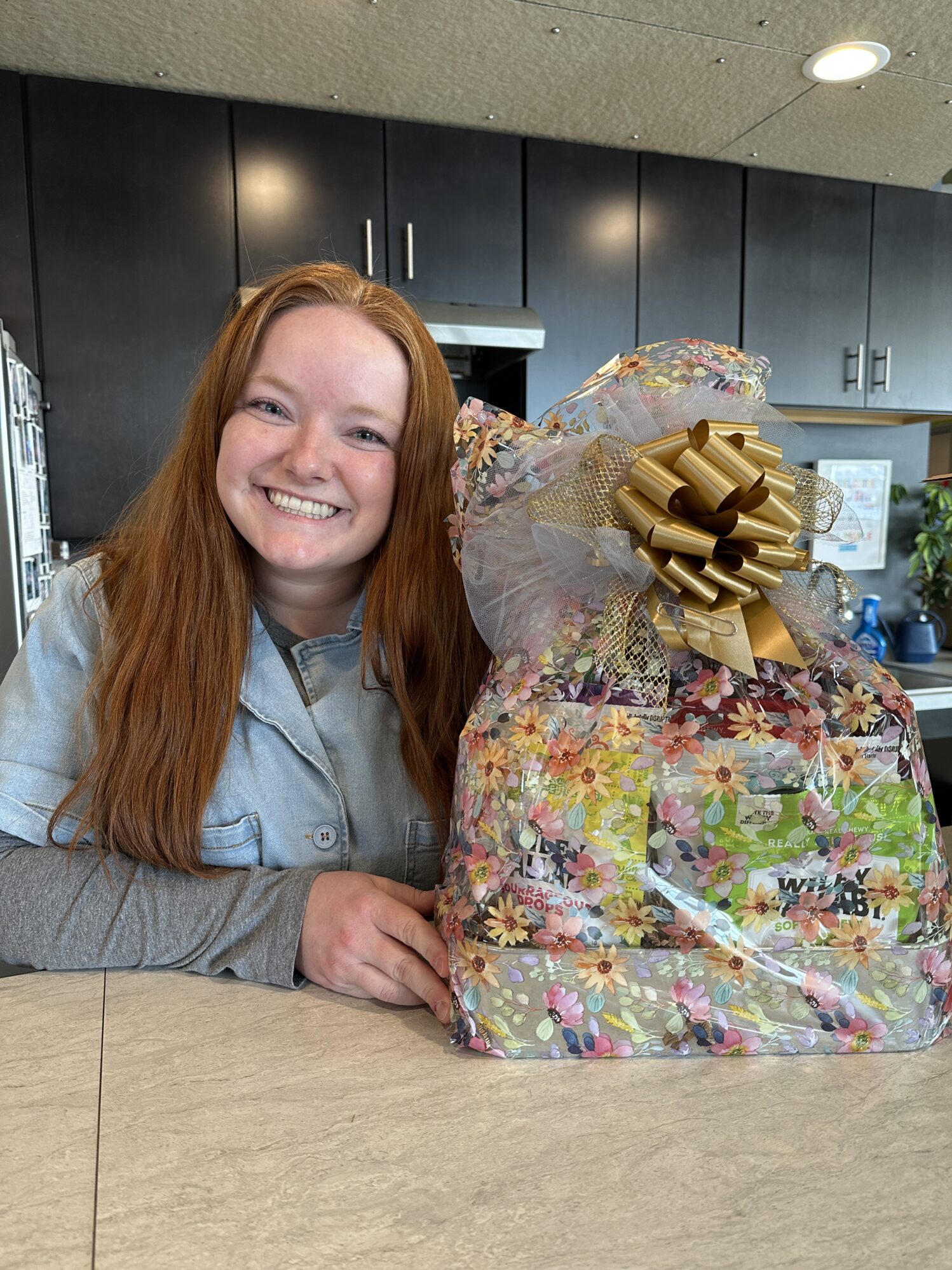 A smiling woman with long red hair and fair skin is sitting at a kitchen counter, posing with a gift basket wrapped in floral-patterned cellophane and adorned with a large gold bow. She is wearing a light blue denim shirt over a gray long-sleeve top. The gift basket contains various items, including snacks and other treats, and is wrapped neatly. The background features dark kitchen cabinets, a stainless steel range hood, and a light-colored ceiling with recessed lighting. The setting appears to be a modern kitchen.