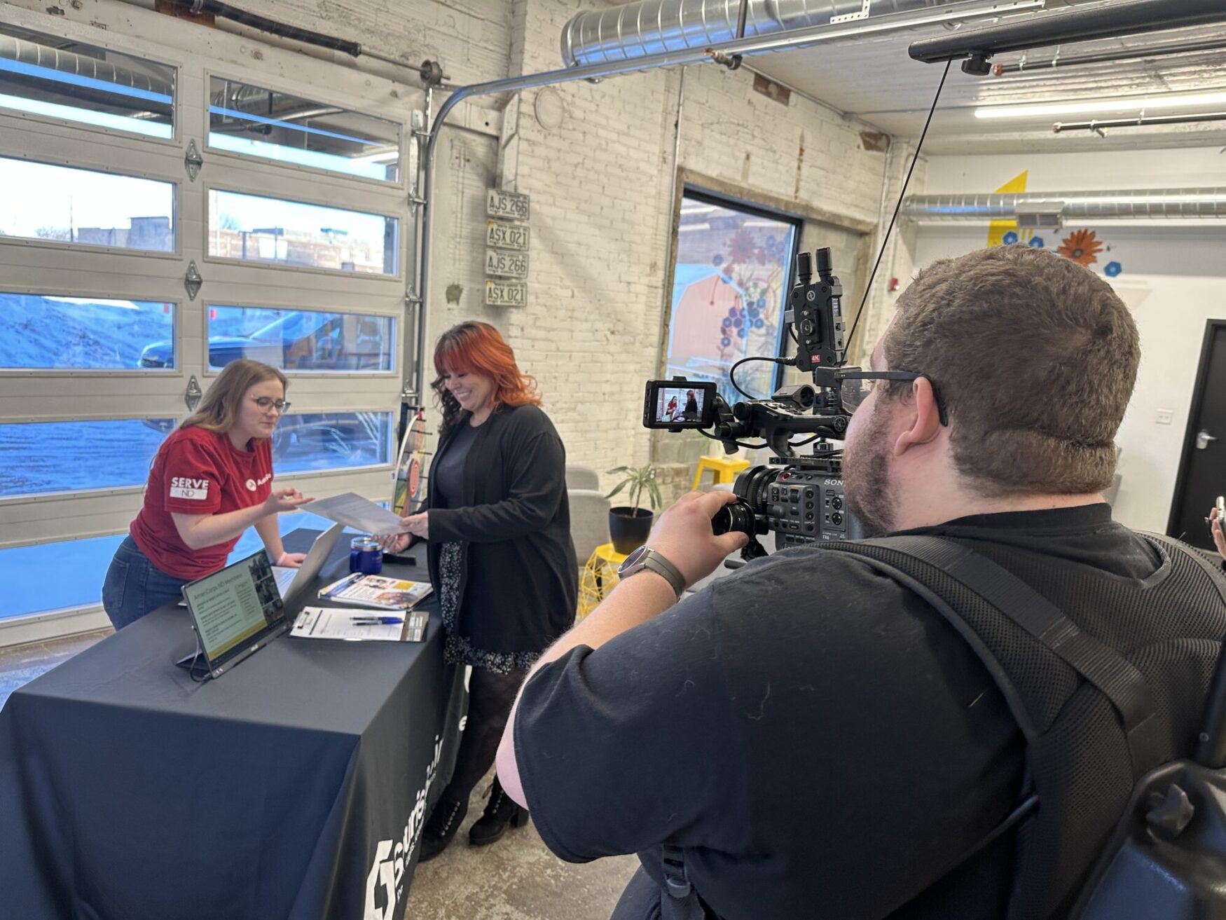 A behind-the-scenes look at a video shoot in an industrial-style space with exposed brick walls and large garage doors. A cameraman wearing a backpack rig films a conversation between two women at an information table covered with a black tablecloth displaying the "Serve ND" logo. One woman, wearing a red "Serve ND" t-shirt, is explaining something while gesturing towards a document, while the other woman, dressed in black, listens and smiles while holding the paper. A laptop, brochures, and other materials are spread across the table. Bright natural light comes through the windows, and colorful artwork decorates the background.