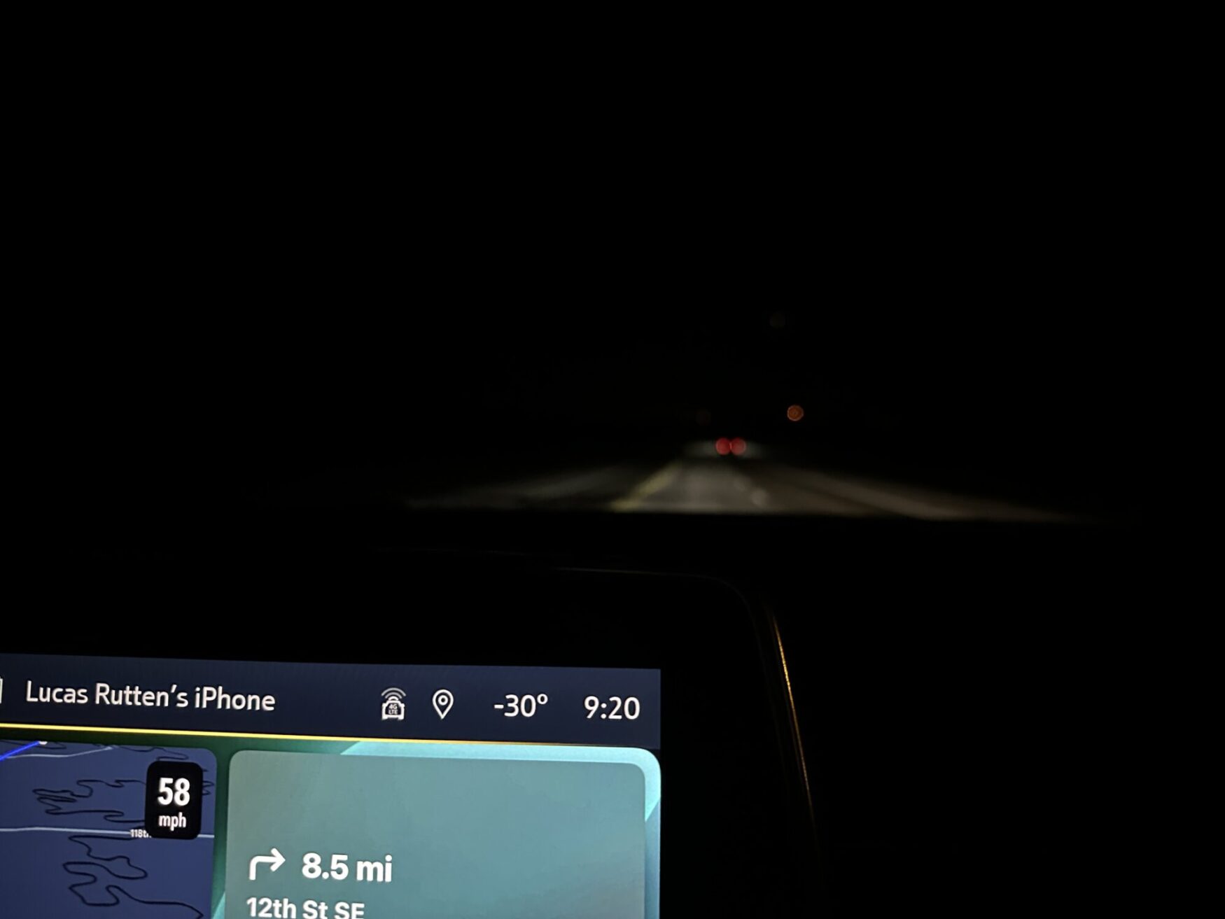 This image shows a nighttime driving scene with a car dashboard displaying navigation directions. The dashboard screen shows the connected device name "Lucas Rutten's iPhone," a current speed of 58 mph, and a navigation instruction indicating 8.5 miles to the next turn onto 12th St SE. The temperature is displayed as -30°, and the time is 9:20. The road ahead is dark, with only a few distant vehicle lights visible.
