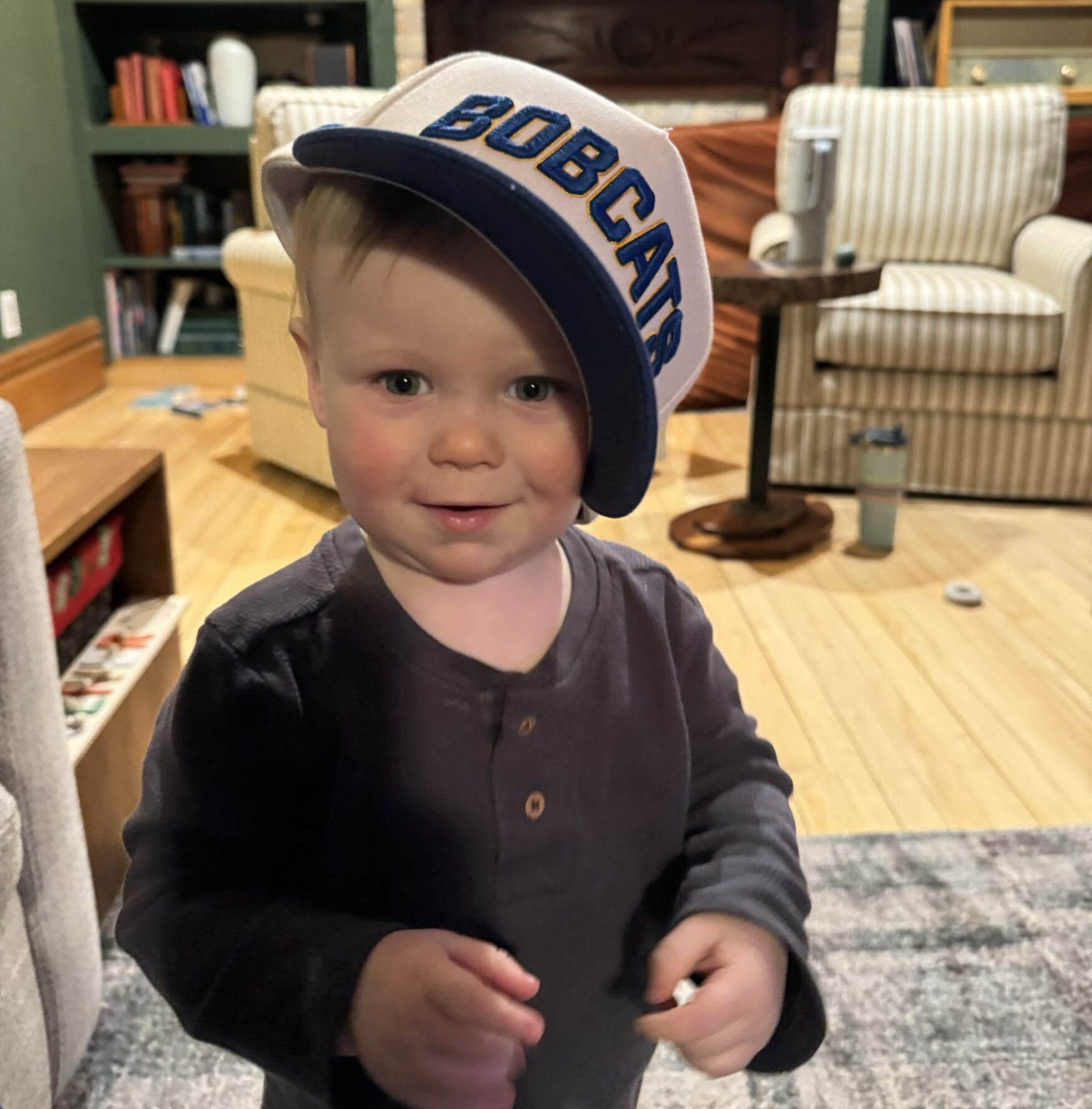 A young toddler with fair skin and rosy cheeks is standing in a cozy living room. The child is wearing a dark long-sleeved shirt and an oversized white and blue baseball cap with the word "BOBCATS" embroidered on it, tilted to one side. The toddler has a slight smile and bright eyes, looking directly at the camera. The background includes a striped armchair, a wooden table, bookshelves, and a soft rug on a hardwood floor, creating a warm and inviting atmosphere.