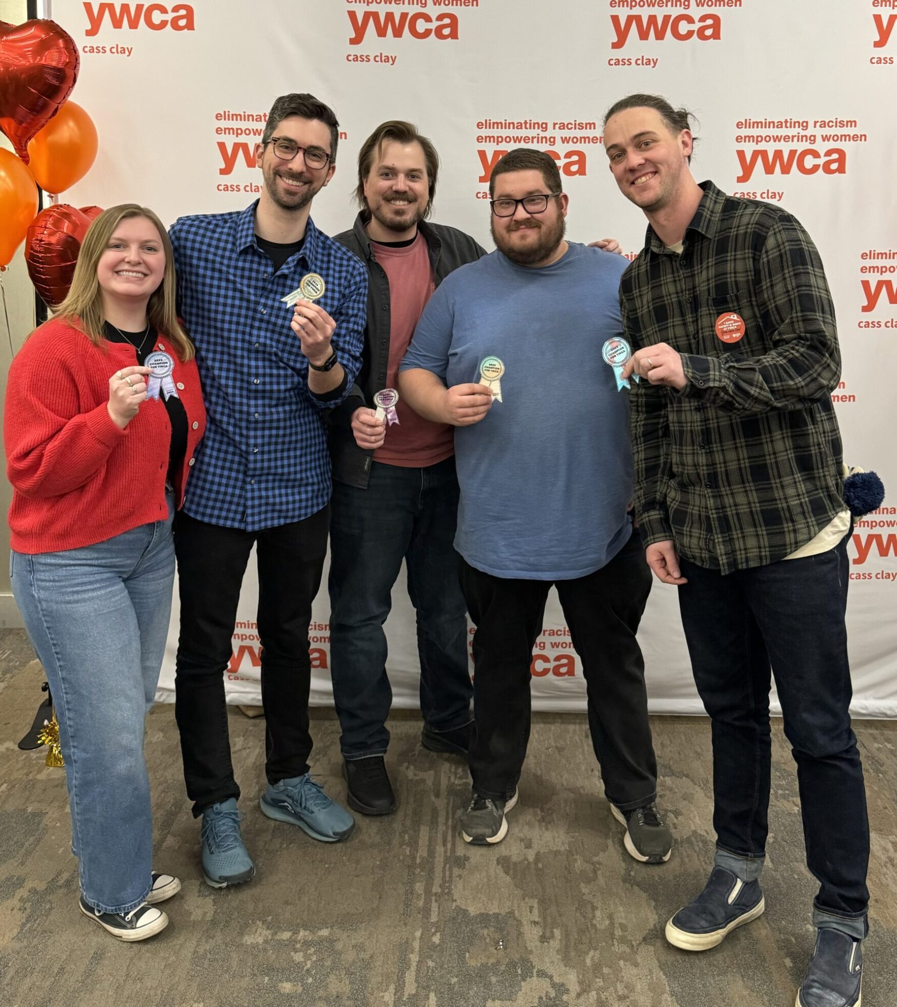 A group of five people, three men and two women, are posing together in front of a backdrop featuring the "YWCA Cass Clay" logo and the phrase "eliminating racism, empowering women." They are all smiling and holding small award ribbons or buttons. The individuals are dressed casually, with a mix of plaid shirts, sweaters, jeans, and sneakers. One person is wearing a red cardigan, while another has a blue and black checkered shirt. There are red and orange balloons to the left side of the image, adding a festive touch. The setting appears to be an indoor event space with a carpeted floor.