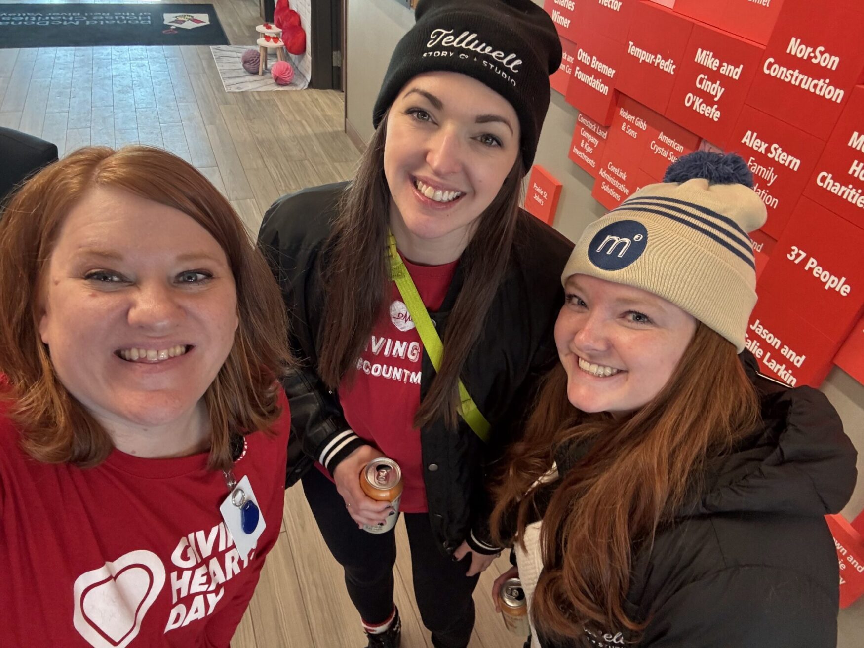 Three women are posing together for a selfie indoors, all smiling warmly. The woman on the left has shoulder-length red hair and is wearing a red "Giving Hearts Day" t-shirt with a heart logo, along with a name badge clipped to her shirt. The woman in the middle has long brown hair, is wearing a black beanie with "Tellwell Story Co. & Studios" embroidered on it, a black jacket, and a red "Giving Hearts Day" shirt underneath. She is holding a canned beverage in her right hand. The woman on the right has long red hair, is wearing a beige beanie with a blue logo, a black puffer jacket, and is smiling brightly. They are standing in front of a wall with red donor plaques featuring various names and organizations, and the floor is made of light-colored wood. The setting appears to be a community or charitable event.