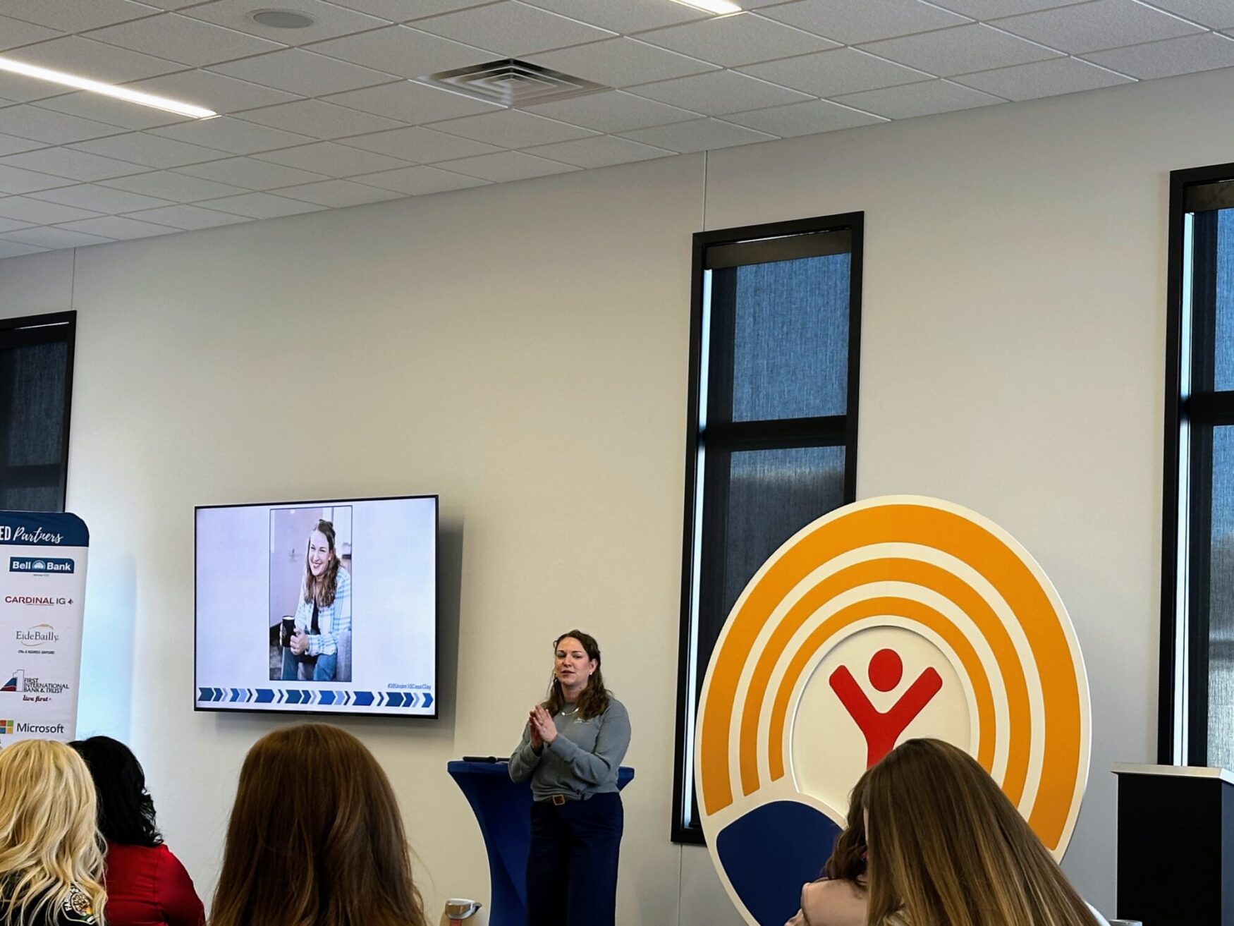 A woman in a gray sweater presents to a seated audience at an indoor event. A large screen behind her displays an image of a smiling woman holding a cup. The venue has modern decor, with a prominent United Way logo in yellow, red, and blue on a stand. Attendees are seated in rows, listening to the speaker.