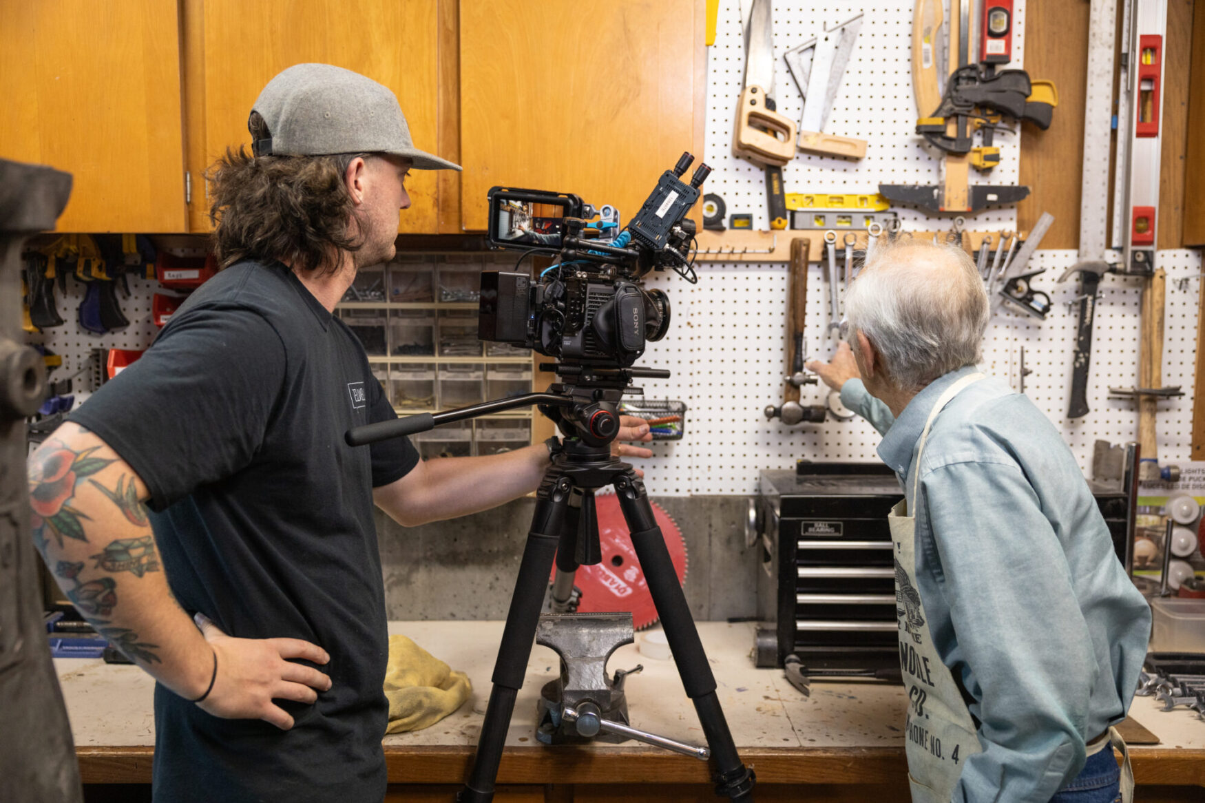 A behind-the-scenes moment in a workshop where a filmmaker and an older craftsman are engaged in a conversation. The filmmaker, a man with long hair, a tattooed arm, and a gray cap worn backward, is wearing a black t-shirt and operating a professional Sony video camera mounted on a tripod. The craftsman, an elderly man with white hair, is wearing a light blue long-sleeved shirt and an apron, pointing to a tool on a pegboard wall filled with neatly arranged hand tools, saws, wrenches, and levels. The setting is a well-organized workshop with wooden cabinets, a workbench, and various tools stored in bins and drawers.