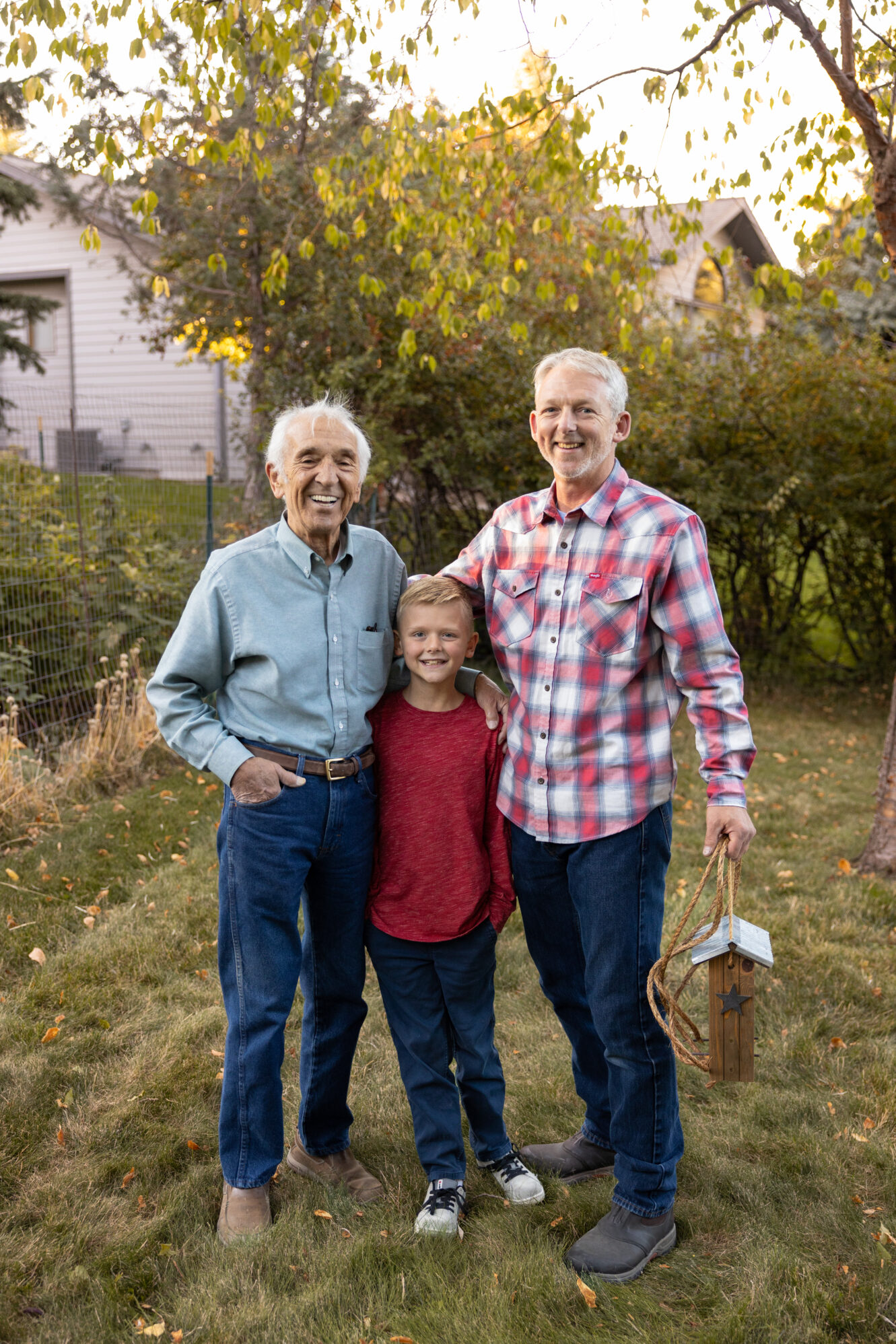 Three generations of a family—an elderly man, a middle-aged man, and a young boy—are standing together in a grassy backyard during what appears to be late afternoon or early evening. The elderly man on the left is wearing a light blue button-up shirt, jeans, and brown shoes, smiling warmly. The young boy in the center is dressed in a red long-sleeved shirt, navy pants, and sneakers, standing happily between the two adults. The middle-aged man on the right is wearing a red and white plaid shirt, jeans, and work boots, holding a rustic wooden lantern with a star design in his left hand. The background features lush trees, a fenced garden, and a house with a steep roof, bathed in golden light from the setting sun.
