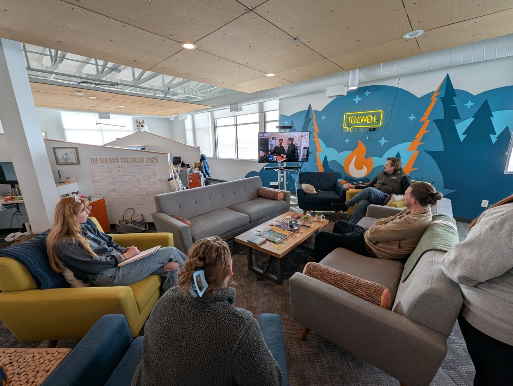 A group of people is gathered in a cozy, modern office lounge, watching a video on a large screen. The room features a vibrant mural with mountains, trees, and a campfire, along with a neon "Tellwell" sign. Various seating options, including sofas and armchairs, are arranged in a semi-circle. A large wall calendar labeled "The Big A** Calendar 2025" is visible in the background. Some individuals are taking notes while others are engaged in discussion. Natural light streams in through the windows, enhancing the collaborative and creative atmosphere.