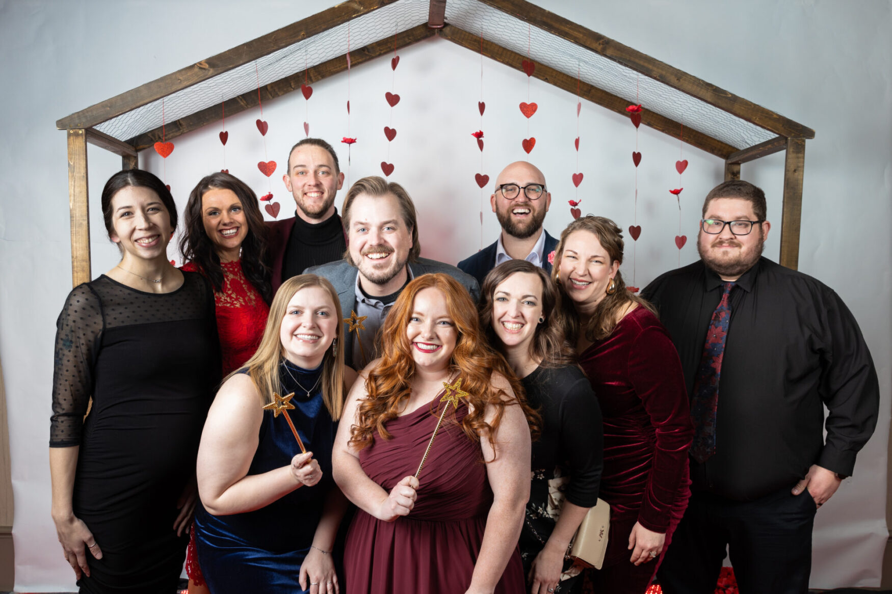 A group of ten people is posing together in a photo booth at a formal event. They are standing under a wooden frame decorated with hanging red hearts and flowers. The group consists of men and women dressed in elegant attire, including black, red, and navy dresses and suits. Two women in the front are holding gold star wands, and they all have cheerful expressions with broad smiles. The background is a white backdrop with the heart decorations adding a festive touch. The setting suggests a Valentine's or sweetheart-themed event, with the participants looking happy and engaged.