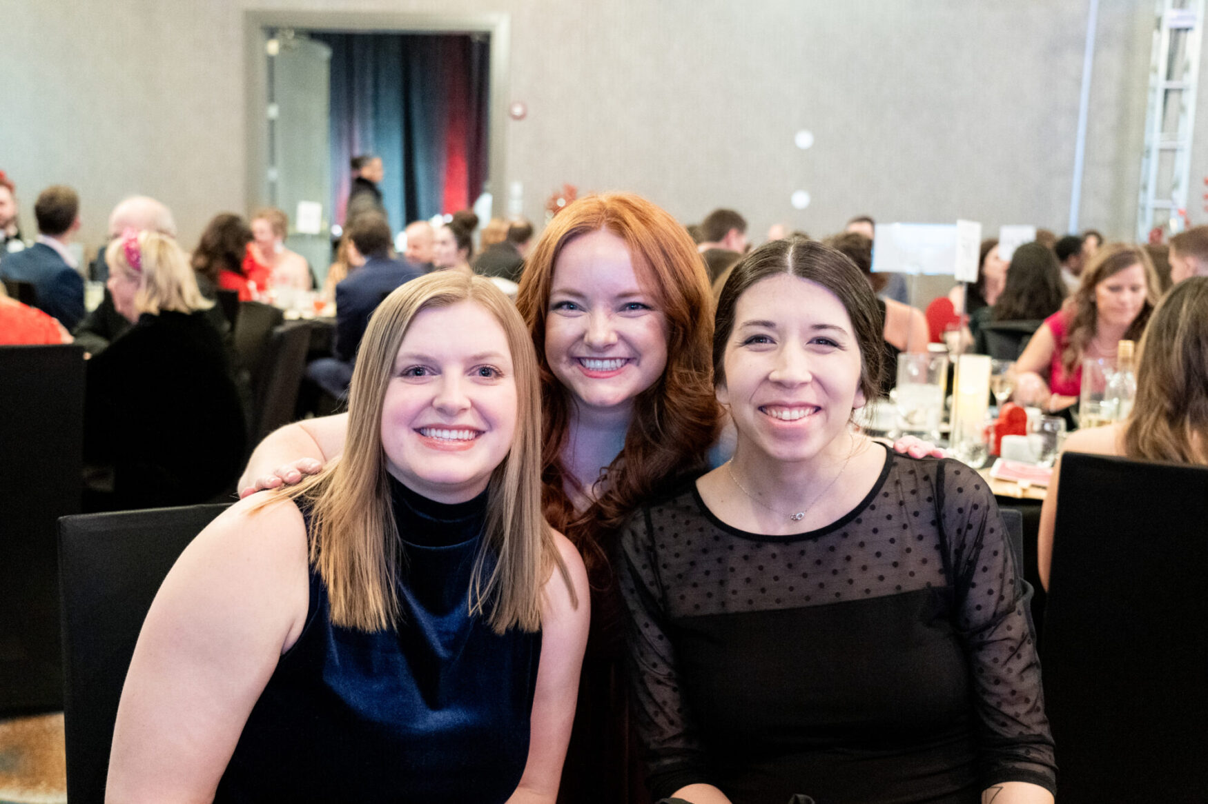 Three women are posing together at a formal event, all smiling warmly at the camera. The woman on the left has straight blonde hair and is wearing a sleeveless dark blue velvet dress. The woman in the center has long, wavy red hair and is leaning in between the other two, dressed in a dark-colored outfit.