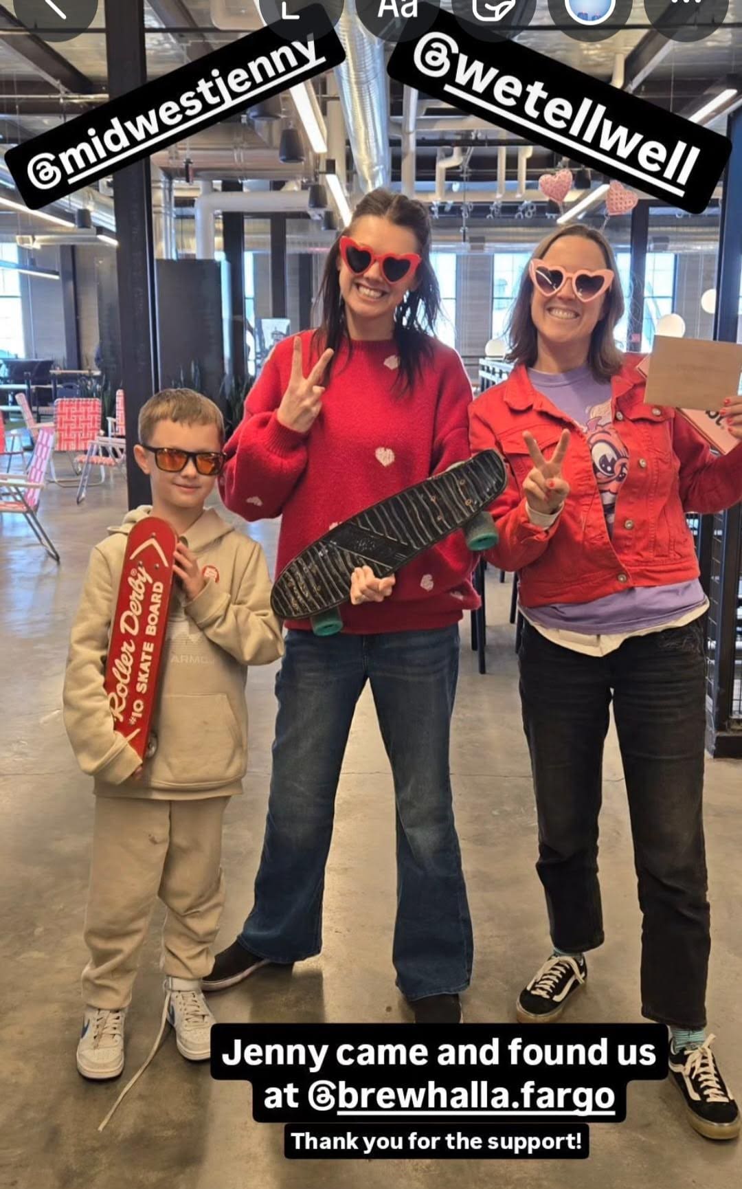 A fun and lively indoor photo of three people—two women and a young boy—posing together in a modern industrial-style space. The women are dressed in red and pink hues and are wearing heart-shaped sunglasses, giving peace signs. The young boy on the left is dressed in a beige hoodie and sweatpants, holding a red skateboard labeled "Roller Derby 10 Skate Board." The woman in the middle is wearing a red sweater with white hearts and is holding a black skateboard. The woman on the right is wearing a red denim jacket over a purple graphic t-shirt, holding an envelope or card. The setting features an open-concept space with large windows, metal beams, and seating in the background. The image has Instagram-style text overlays tagging "@midwestjenny" and "@wetellwell," along with a caption mentioning that Jenny found them at "@brewhalla.fargo" and thanking them for their support.
