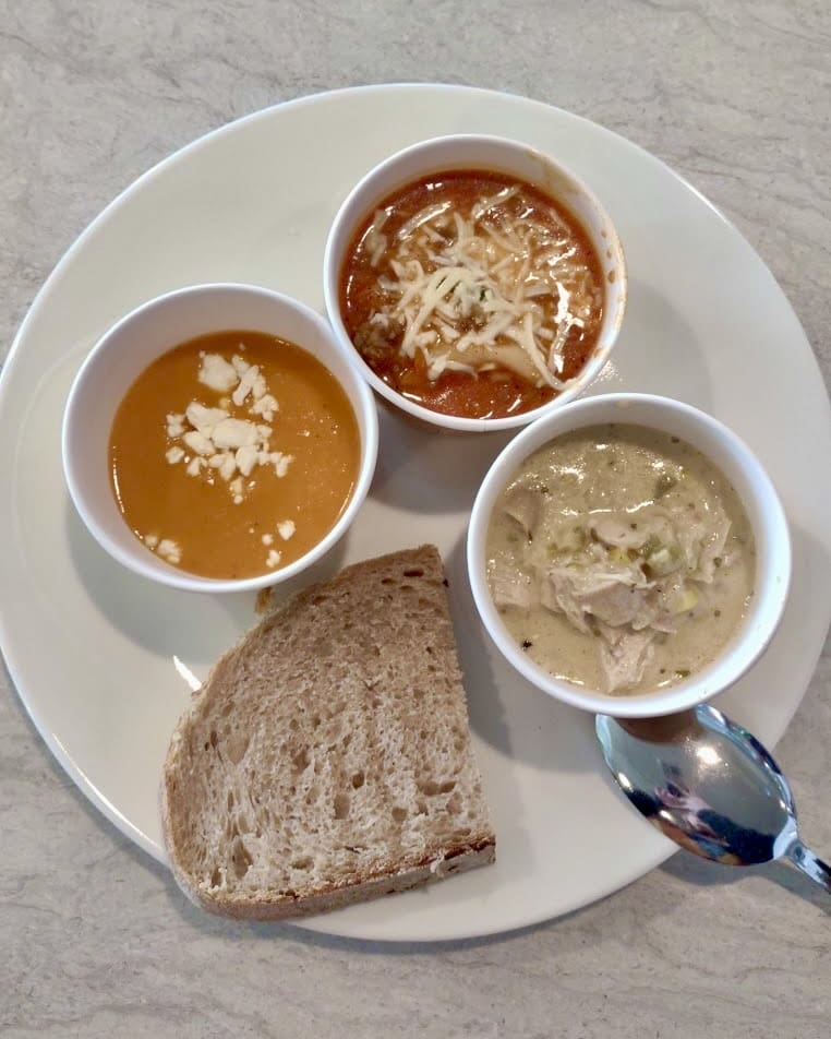 A top-down view of a white plate holding three small bowls of different soups and a slice of rustic bread. The soup in the top-right bowl is a tomato-based soup with melted shredded cheese on top. The bottom-right bowl contains a creamy, white soup with shredded chicken and herbs. The bottom-left bowl features an orange-colored soup, likely butternut squash or sweet potato, topped with crumbled cheese. A metal spoon rests on the right side of the plate, reflecting light. The plate is placed on a light-colored countertop.