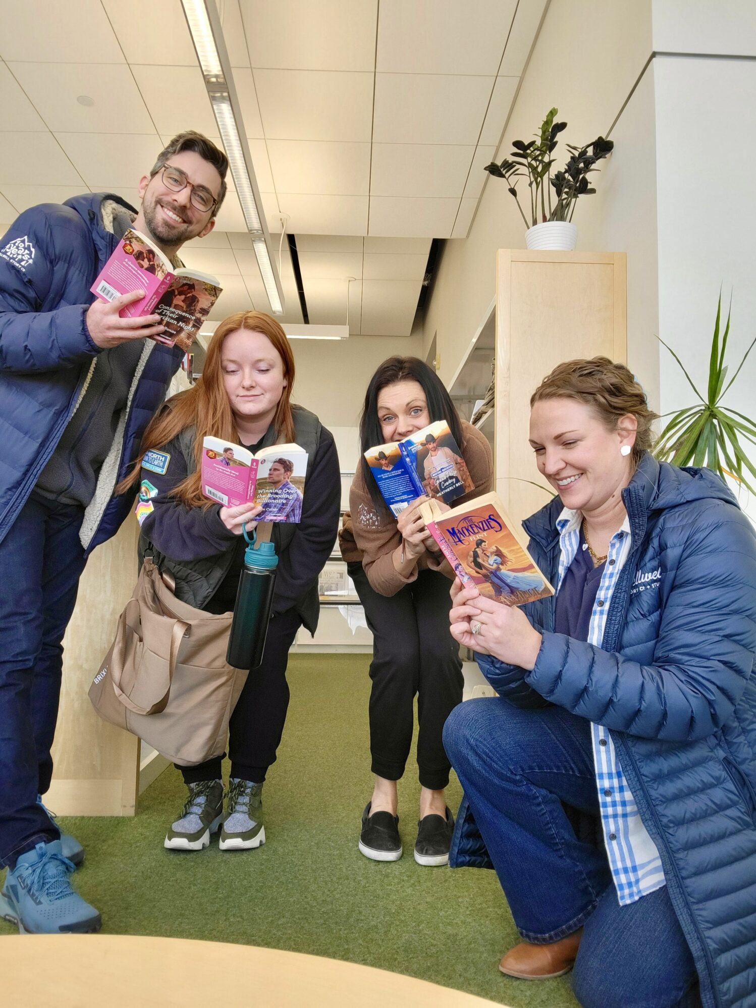 Four coworkers pose together in an office space with green carpeting and wooden shelving. They are all holding and reading romance novels with dramatic covers, smiling and laughing. The man on the left, wearing glasses and a blue jacket, holds his book up excitedly. The woman in the center-left, with long red hair and a gray vest, looks down at her book with a serious expression. The woman in the center-right, dressed in black, peeks over her book playfully. The woman on the far right, wearing a blue puffer jacket and plaid shirt, kneels while grinning at her book. The background includes potted plants and office furniture.