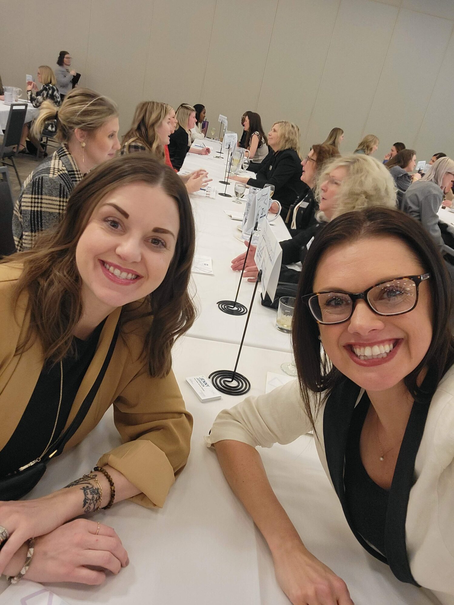 A lively networking event with two smiling women in the foreground taking a selfie. They are seated at a long table, dressed in business casual attire. The background features a room full of other women engaged in conversations, suggesting a professional or networking setting.