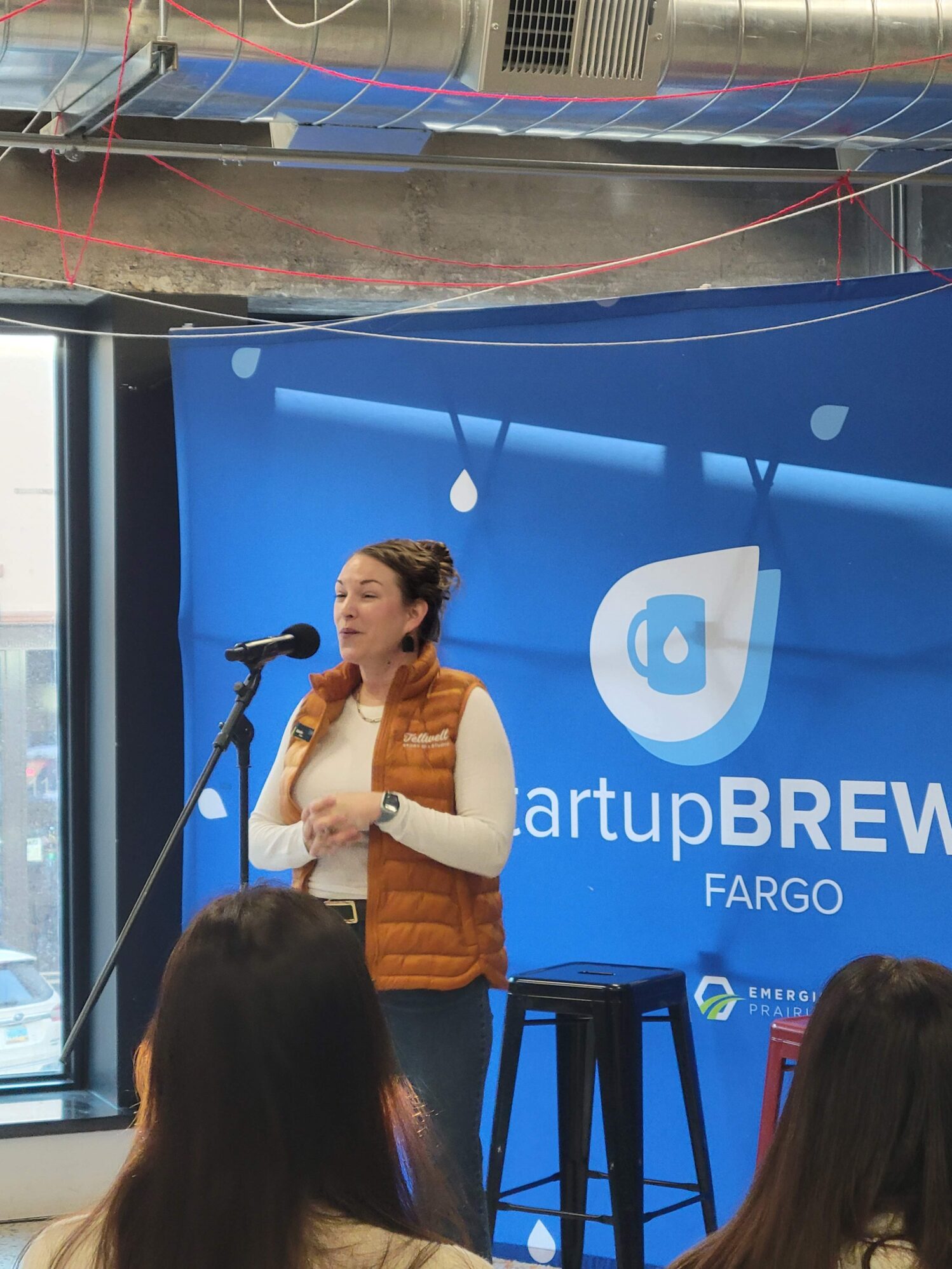 A woman stands in front of a StartupBrew Fargo backdrop. She is wearing a white turtleneck with a Tellwell Story Co. orange puffer vest. She is standing at the front of the room, speaking into a microphone.