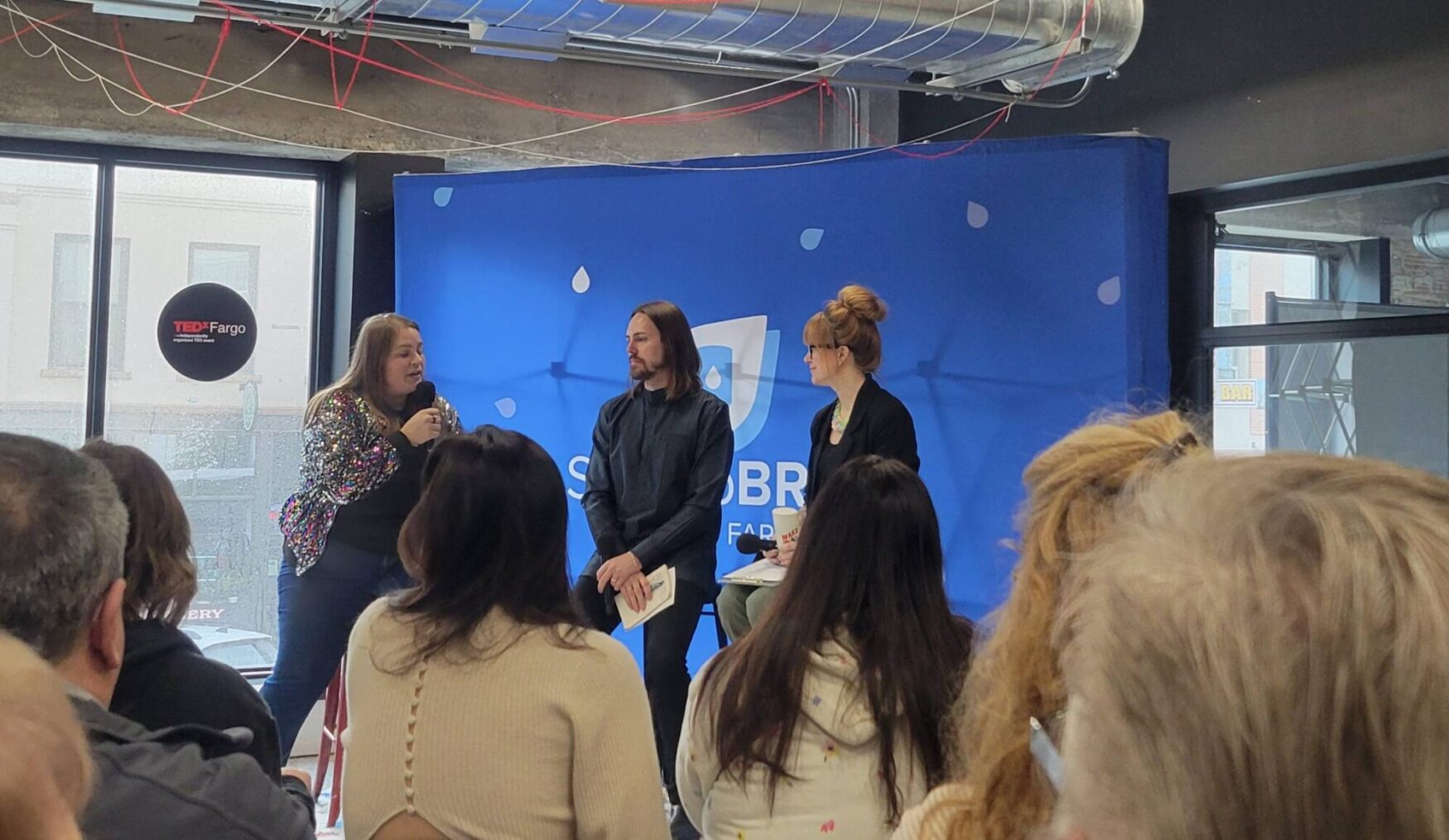 A panel discussion at a TEDxFargo event, featuring three speakers on stage. A woman in a sequined jacket holds a microphone while speaking, while a man with shoulder-length hair and another woman with a topknot listen attentively. The backdrop features a blue banner with a logo and raindrop designs. The audience, seen from behind, watches the discussion in a modern, industrial-style venue with exposed ductwork and large windows.