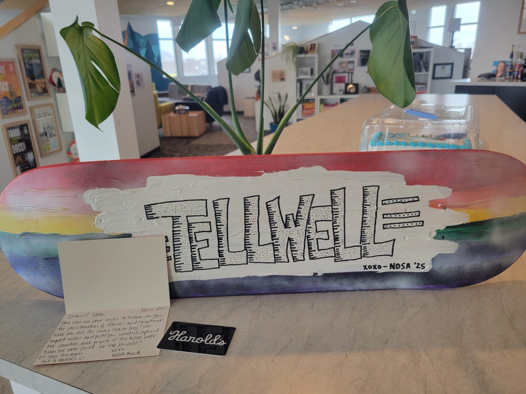 A painted skateboard is propped up on a plant. This skateboard is rainbow colored with Tellwell painted on it in black and white. There is a letter written next to it and a gift card to Harold's.