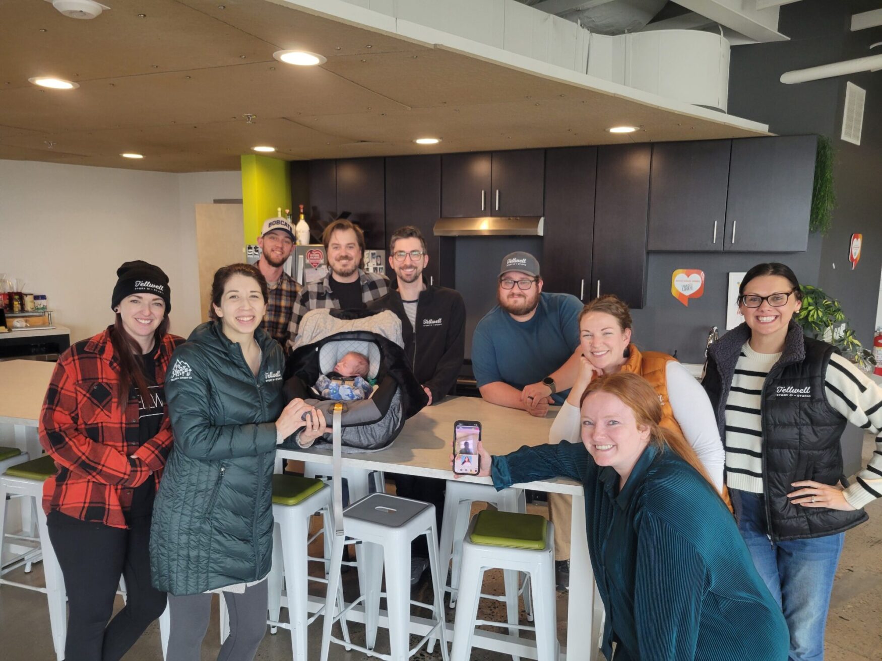 This image shows a group of ten people, including a baby in a car seat, gathered in a modern kitchen space with black cabinets and a green accent wall. Some individuals are wearing branded clothing with the name "Fidwell." A woman in the foreground, dressed in teal, is crouching with a smartphone displaying a photo, smiling at the camera. Others in the group are standing and posing cheerfully, with some wearing flannel or vests. The setting has white stools around a light-colored countertop, and decorative heart-shaped signs are visible on the wall.