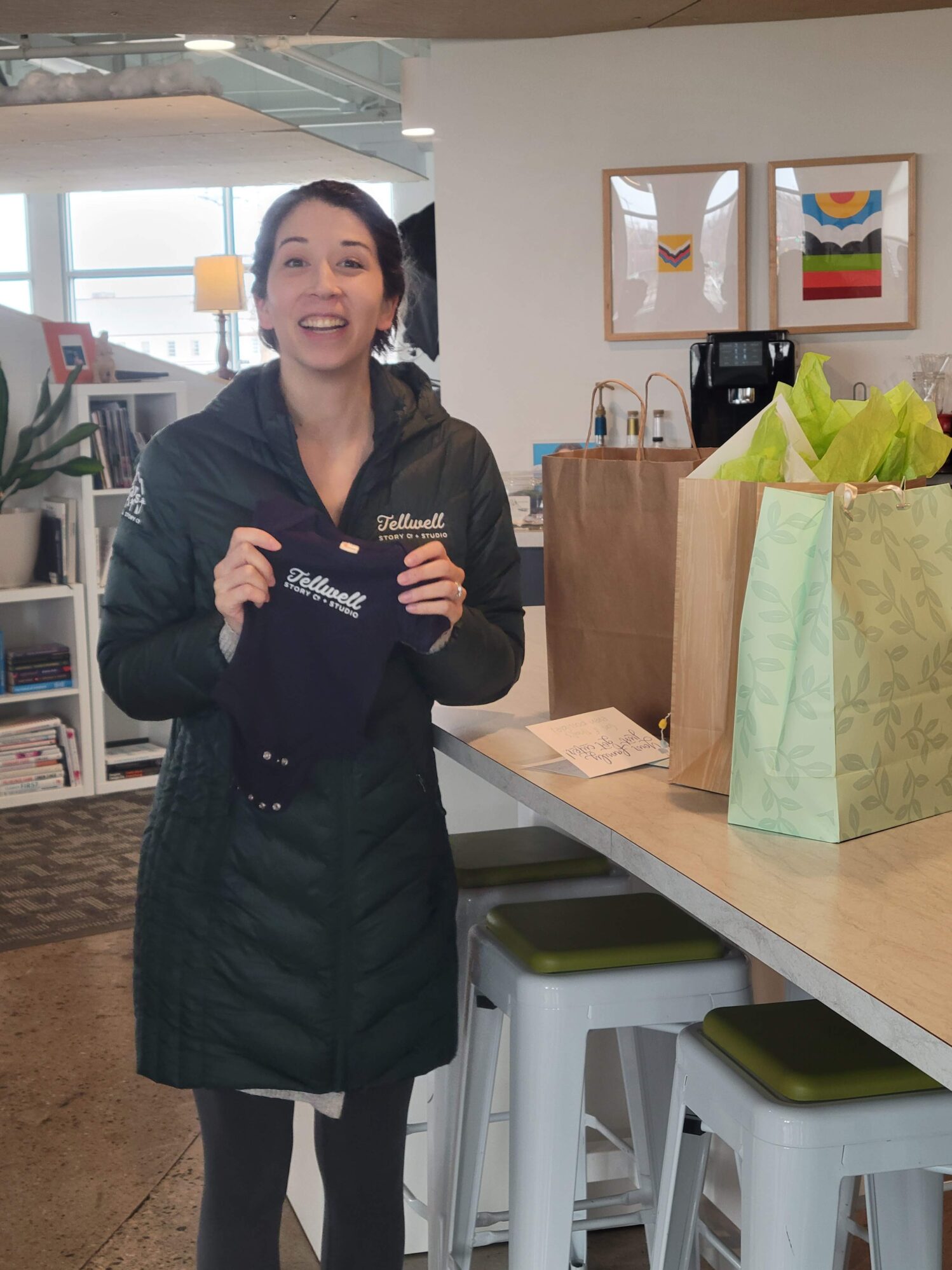 A woman wearing a Tellwell Story Co. winter jacket stands in an office setting holding up a navy blue baby onesie that also ways Tellwell Story Co.