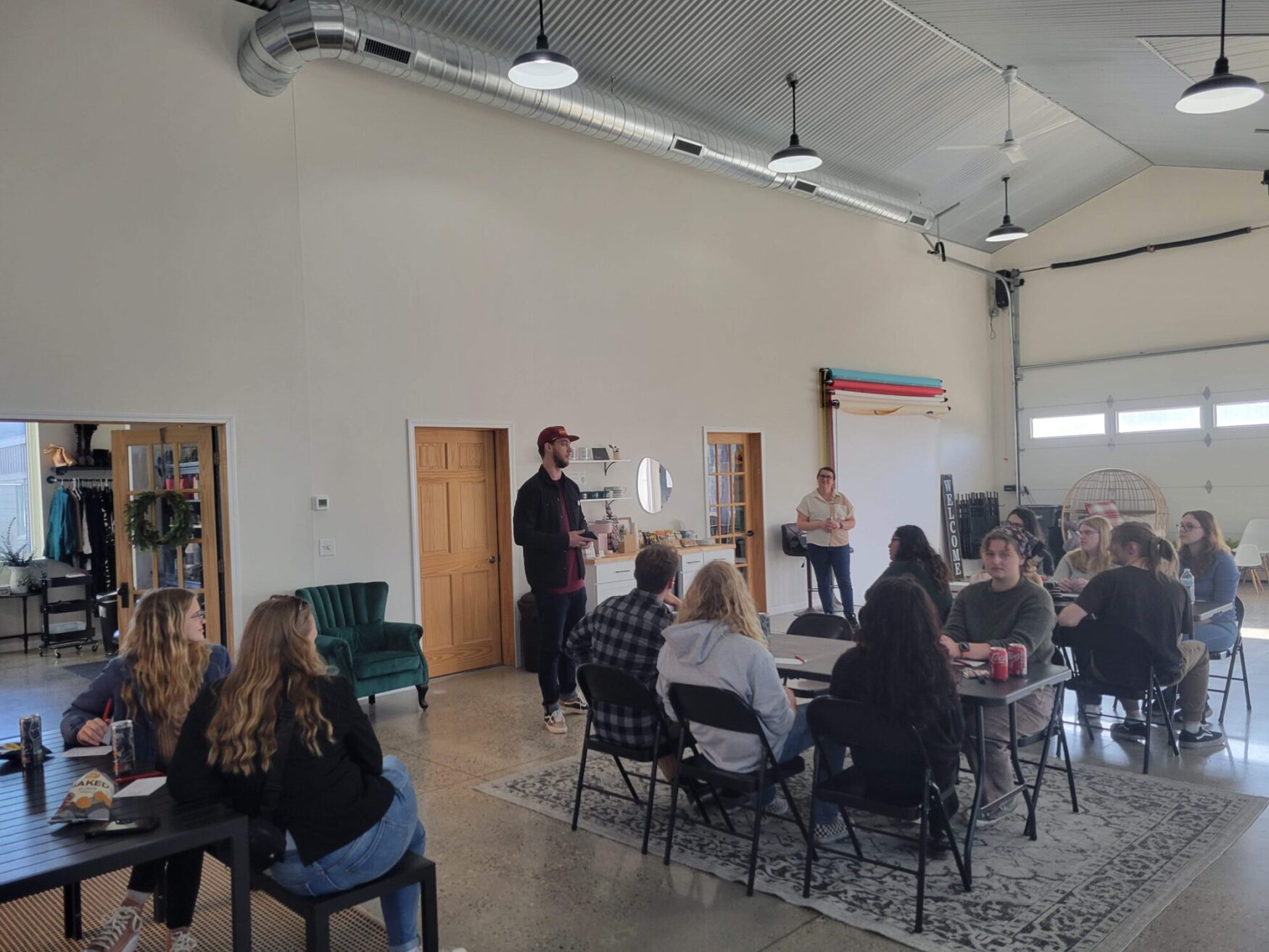 This image shows a spacious, industrial-style room with high ceilings, large garage doors, and exposed ductwork. A group of people is seated at black tables arranged in a semi-circle, attentively listening to a man in a red cap and dark jacket who is standing and speaking. A woman in a white shirt and glasses stands nearby holding a notebook. The walls are decorated with colorful rolled-up backdrops, a "Welcome" sign, and a mirror. On the left, a small boutique-like area with clothing and a wreath is visible. The setting has a casual and collaborative atmosphere, with attendees taking notes and drinks on the tables.