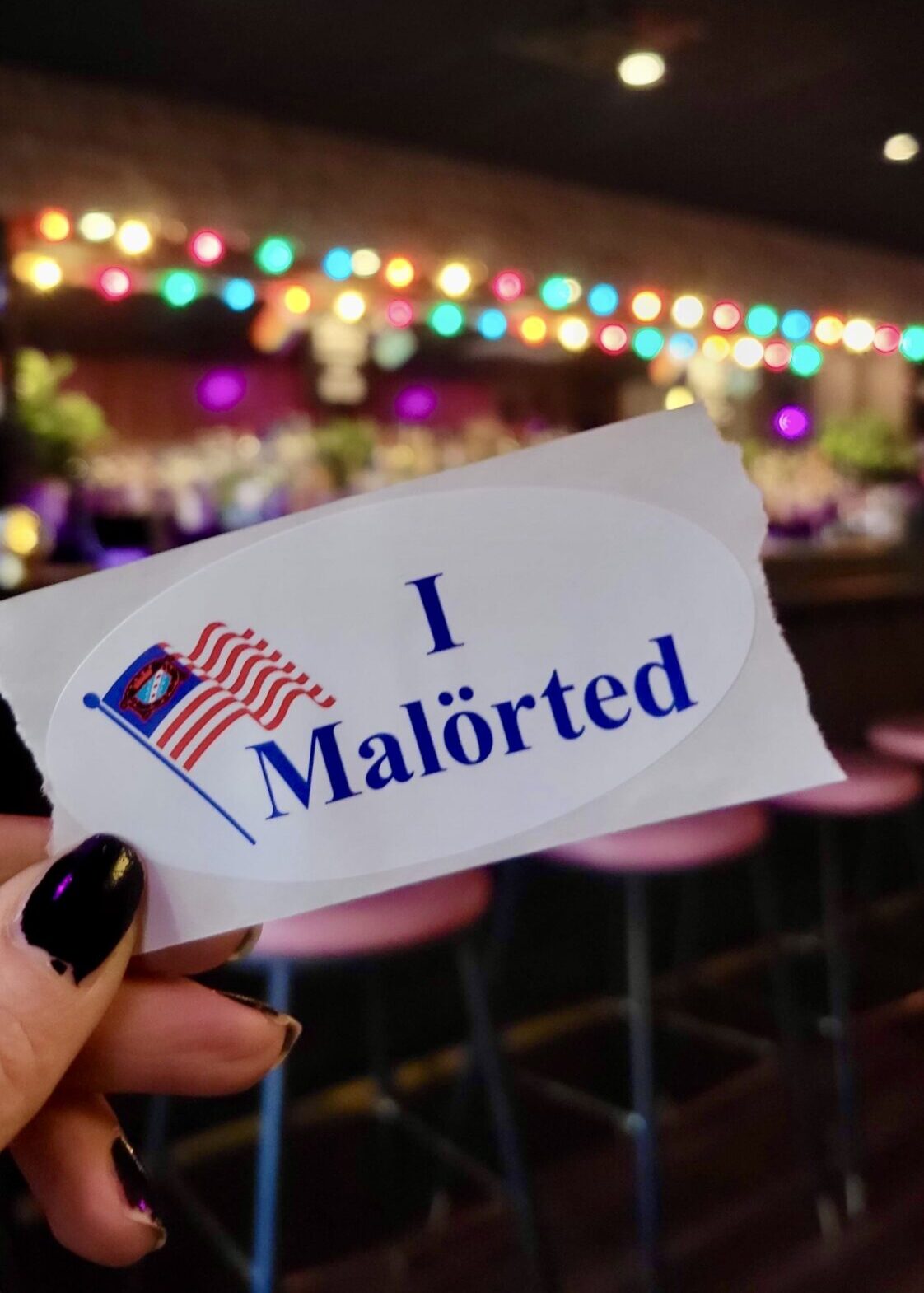 A hand with black-painted nails holds a white sticker that mimics an "I Voted" sticker but reads "I Malörted," featuring a waving flag with a Chicago flag design. The background is blurred, showing a dimly lit bar with colorful string lights, pink barstools, and plants, creating a lively and cozy atmosphere.