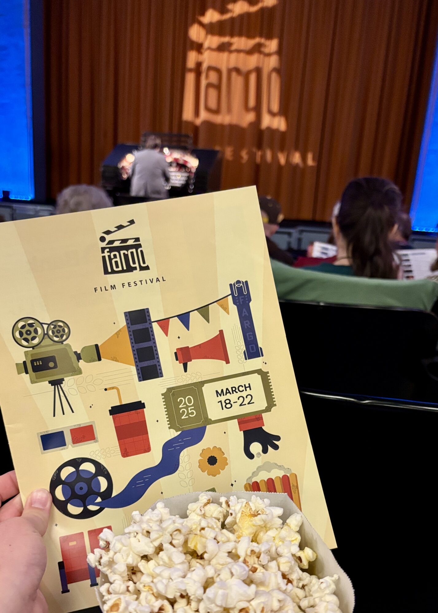 A person holds a Fargo Film Festival program and a container of popcorn inside a theater. The program cover features colorful illustrations of film-related items such as a vintage camera, film reels, a megaphone, and a movie ticket with the festival dates: March 18-22, 2025. In the background, the theater stage is visible with a large "Fargo Film Festival" logo projected onto a curtain. A person is playing a pipe organ, and audience members are seated, some holding similar programs.