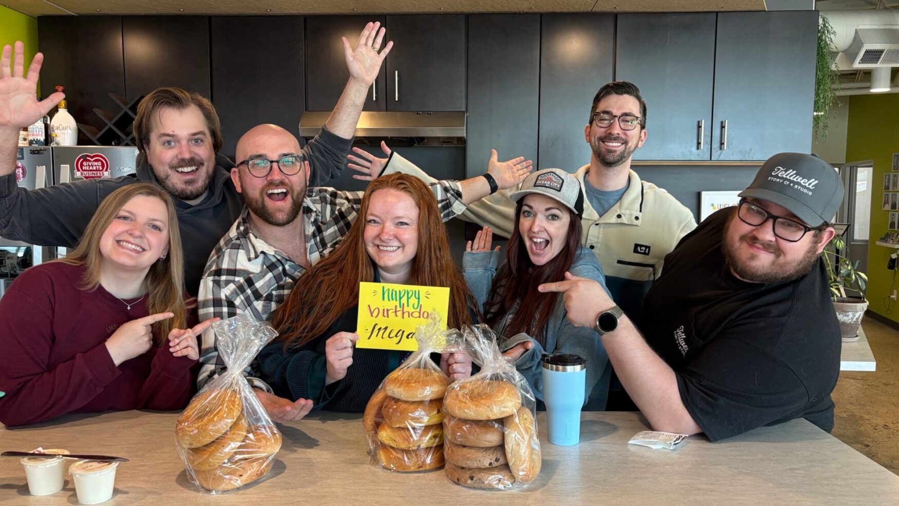 A group of cheerful coworkers gathers around a kitchen counter, celebrating a birthday. The birthday person, a woman with red hair, holds a handmade sign that says "Happy Birthday Megan" in bright, colorful letters. Everyone is smiling, pointing at her, and striking playful poses. In front of them, two large bags of assorted bagels and small containers of cream cheese sit on the counter. The setting is a modern office kitchen with dark cabinets, a stainless steel fridge, and a lively atmosphere.