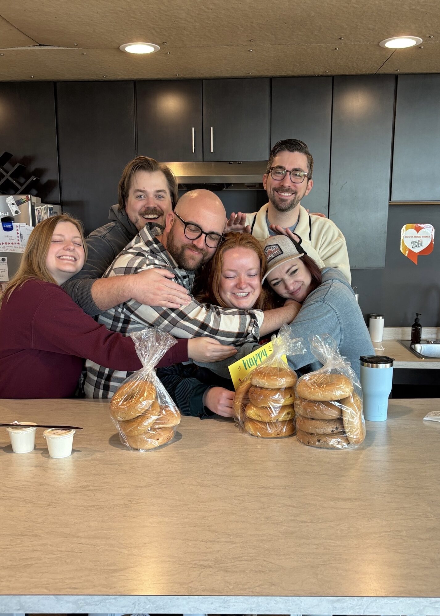 A group of six friends is gathered in a kitchen, sharing a big, joyful group hug. They’re all smiling and laughing, some with their eyes closed, clearly enjoying the warm moment. The group includes a mix of people wearing casual, cozy clothes — one person is even wearing a hat. In front of them on the counter, there are several bags of bagels and a couple of small containers of cream cheese. There’s also a yellow card tucked among the bagels that says "Happy." The kitchen has dark cabinets, and you can spot a coffee maker and a couple of travel mugs on the counter in the background. The vibe feels like a close-knit group having a laid-back, happy gathering.