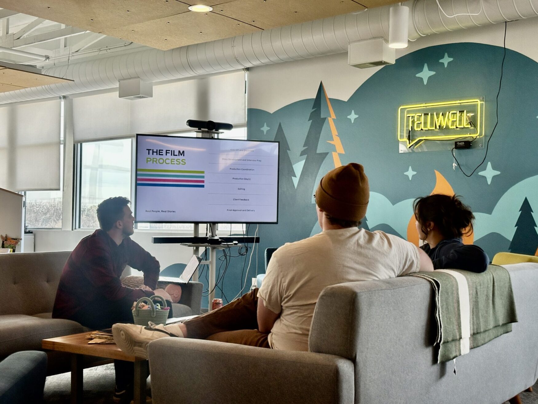 A group of people sits on couches in a creative office space, watching a presentation on a large screen titled "THE FILM PROCESS." The screen outlines different stages of filmmaking. A neon sign on the wall reads "TELLWELL," and the room features a colorful mural with trees and mountains. The setting has a casual, collaborative atmosphere.