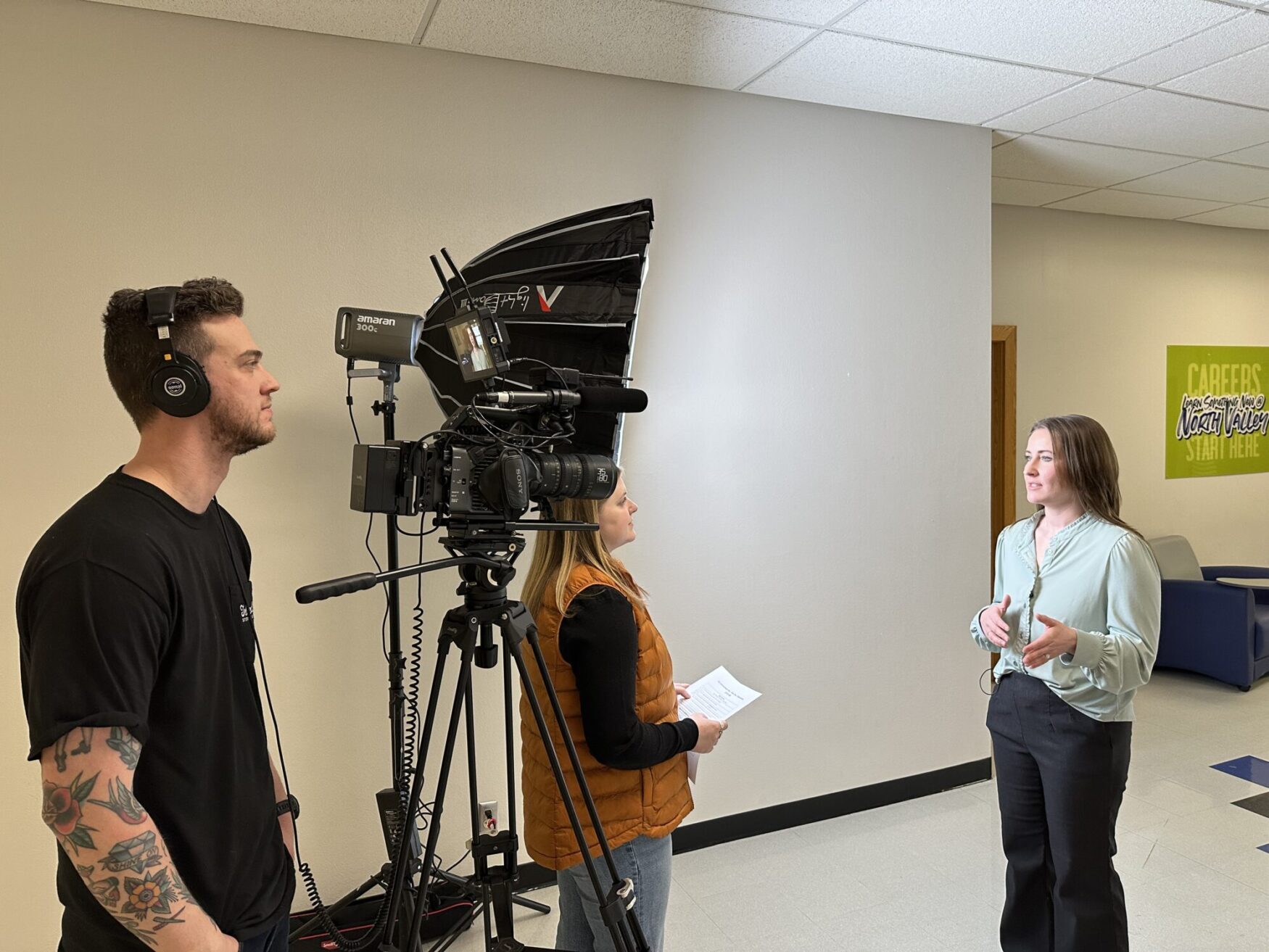 A small video production team is filming an interview in a professional indoor setting. A woman in a light green blouse and dark pants is speaking and gesturing with her hands, wearing a small microphone. The interviewer, wearing an orange vest and holding a sheet of paper, is attentively listening. A cameraman with tattoos and headphones is operating a professional video camera mounted on a tripod, with additional lighting equipment set up nearby. A green sign on the wall in the background reads "CAREERS" and mentions North Valley, suggesting the location may be related to education or job opportunities.