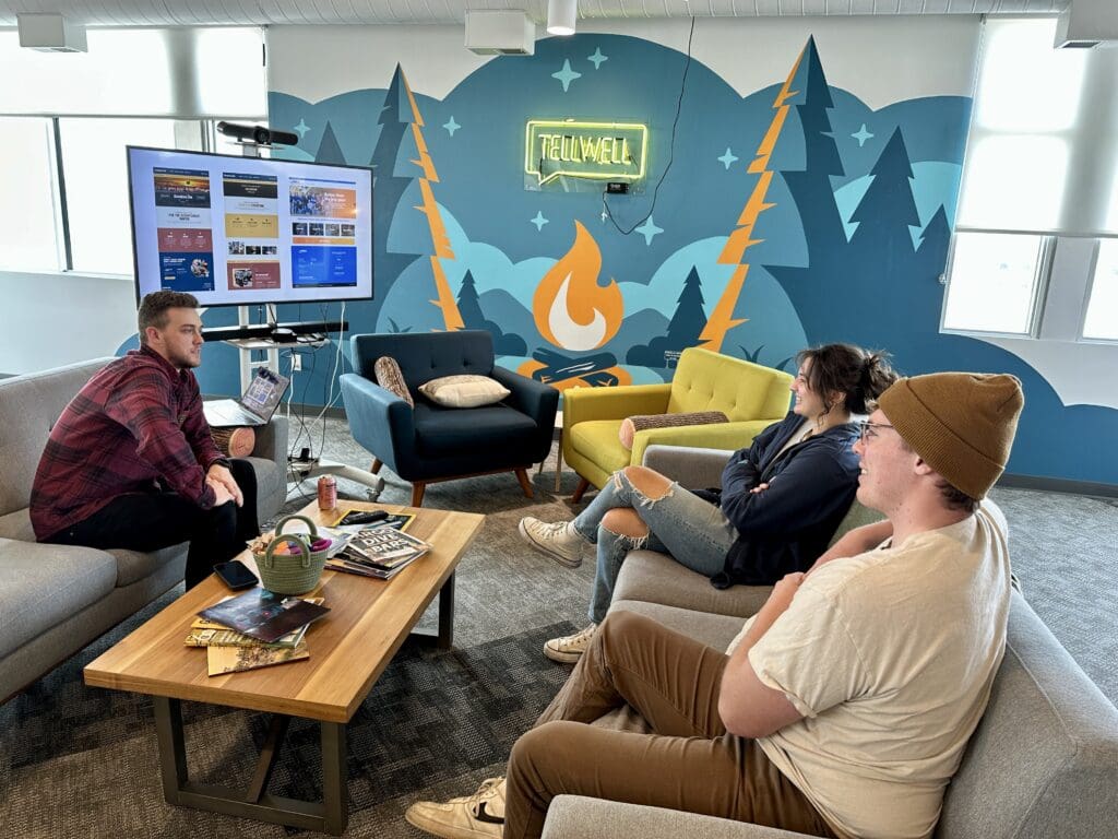 Three people sit in a colorful lounge with a camping-themed mural and a neon "TELLWELL" sign. They relax on modern furniture, engaged in conversation. A man in a red plaid shirt speaks while the others, one in a hoodie and another in a beanie, listen. A TV screen displays a website layout, with a laptop and soda can nearby. The coffee table holds magazines and a small basket, enhancing the laid-back vibe.