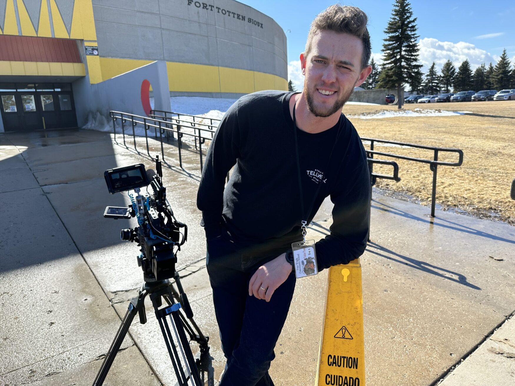 A man stands outside a concrete building, leaning on a traffic cone next to a camera and tripod set-up. The sun is shining bright, causing him to squint while he smiles.