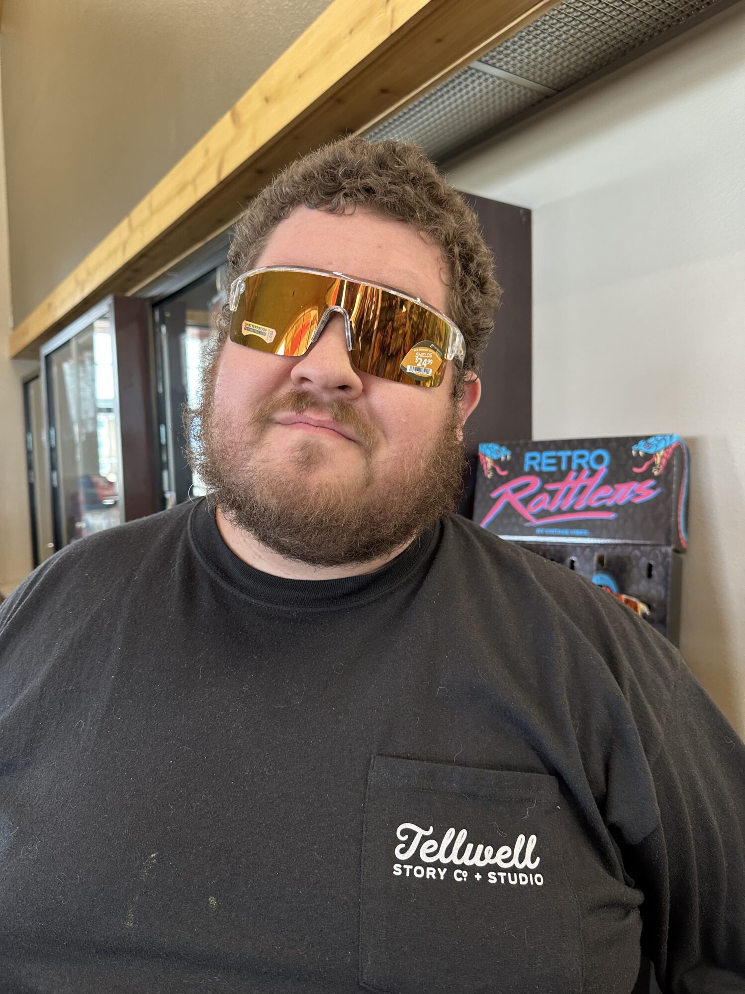 A man wearing a Tellwell Story Co. shirt stands in a convenience story wearing funny sunglasses.