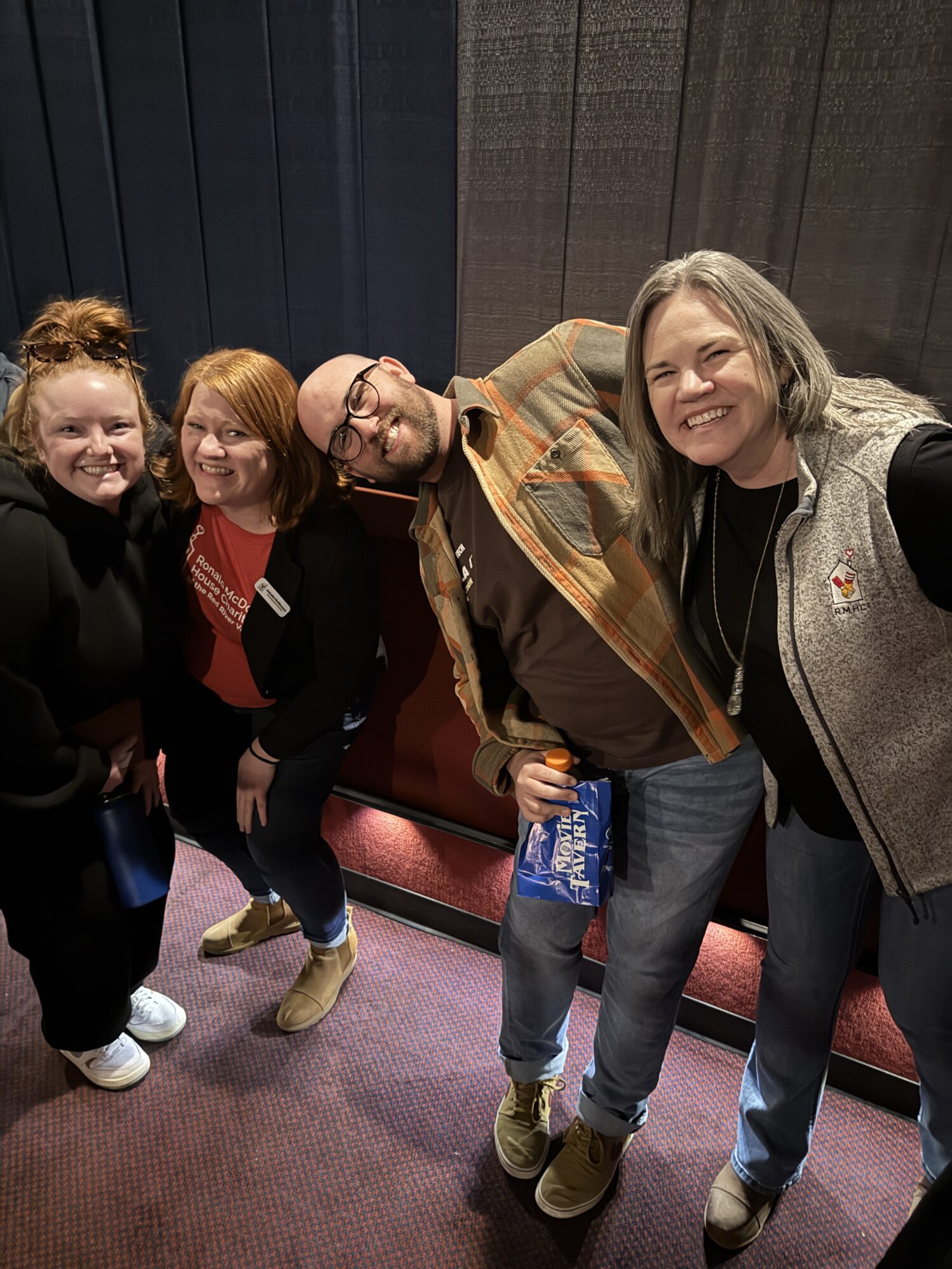 This image shows four people posing together indoors, standing on a red carpeted floor with dark curtains in the background. They are smiling and leaning towards each other playfully. One person, a bald man wearing glasses and a brown shirt with a plaid jacket, is holding a bag of popcorn and a drink. The others are dressed casually, with one wearing a vest featuring the Ronald McDonald House Charities logo. The atmosphere appears cheerful and relaxed.
