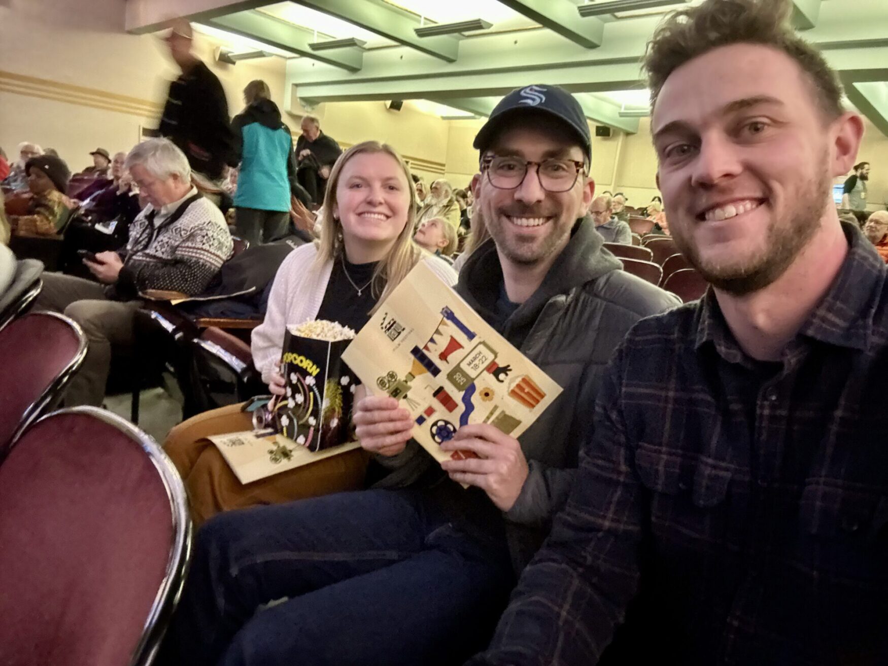 Three people sit together in a theater, smiling at the camera. The person on the left is a woman with blonde hair, wearing a white sweater, holding a box of popcorn and a program. The person in the middle is a man with glasses, wearing a black cap with an "S" on it, a dark hoodie, and a black puffer jacket, also holding a program. The person on the right is a man with short, curly brown hair, wearing a dark plaid shirt, taking the selfie. The background shows other theatergoers settling into their seats, with some standing and chatting.