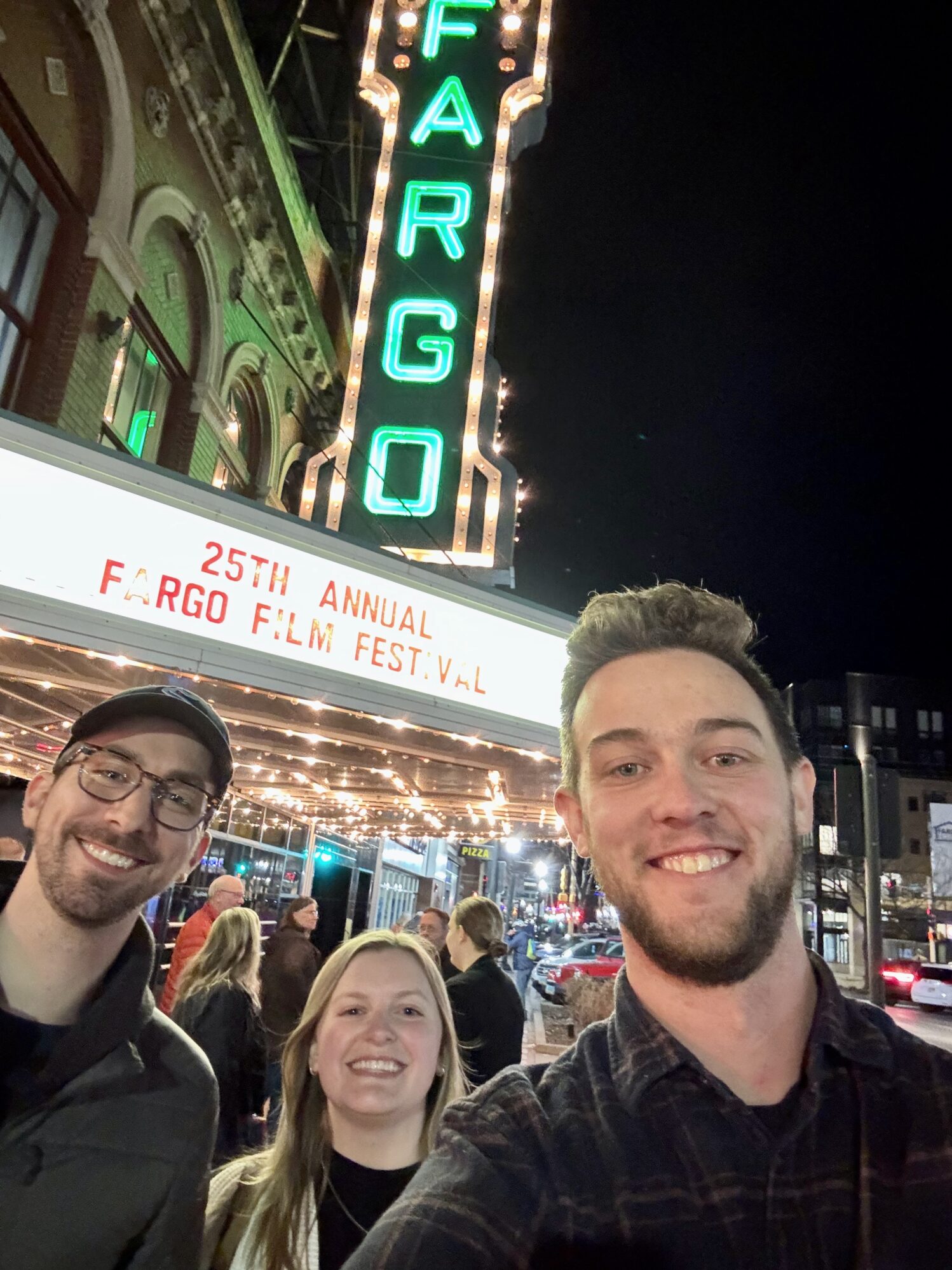 Three people smile for a selfie in front of the Fargo Theatre at night. The theater's iconic neon sign glows above them, and the marquee reads "25th Annual Fargo Film Festival." The person on the right, wearing a dark plaid shirt, holds the camera. The person on the left wears glasses, a black cap, and a dark jacket, while the person in the middle has blonde hair and is wearing a white sweater. The background is bustling with festival attendees and city lights.