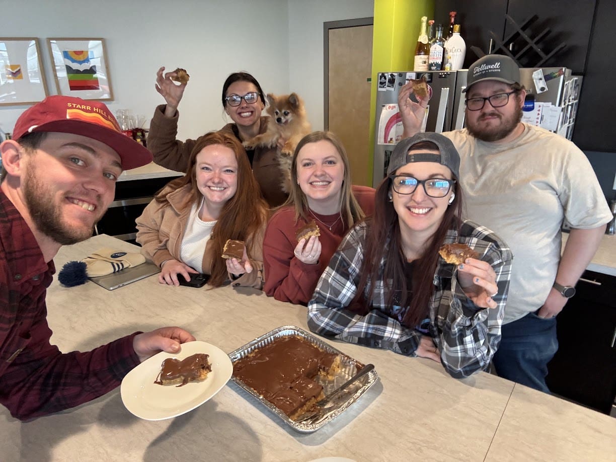 A group of six friends, including a small dog, are gathered around a kitchen counter, smiling and posing for a photo while holding pieces of dessert. The man in the foreground, wearing a red cap and plaid shirt, is holding a plate with a serving of the treat. A foil tray of the dessert, partially eaten and covered in chocolate, is on the counter. The kitchen has a modern style with a green accent wall, artwork, and various bottles on a shelf. The group is casually dressed, with one woman wearing a plaid shirt and a backward cap, and another man in a gray shirt and baseball cap. The atmosphere is cheerful and relaxed.