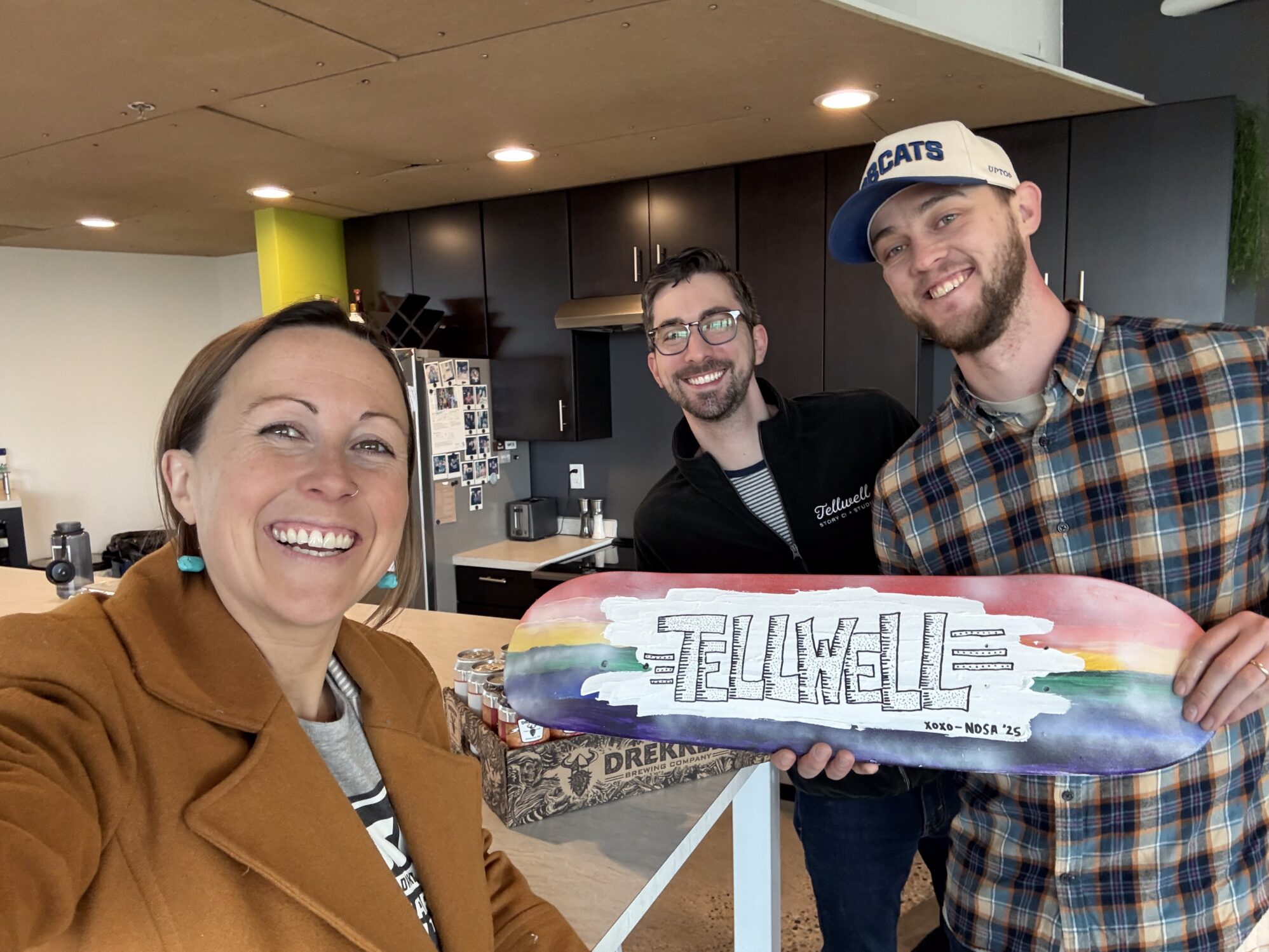 A woman stands with two men holding a customized skateboard. This board is painted in rainbow colors with the word Tellwell painted on it in black and white.