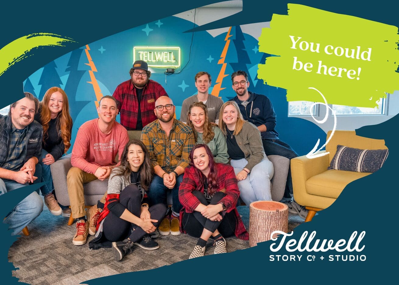 A group of smiling people poses in a cozy, colorful office space with a mural of trees and a neon "Tellwell" sign on the wall. A yellow chair is empty with a playful green text bubble above it reading, "You could be here!" accompanied by a white arrow pointing to the seat. The Tellwell Story Co. + Studio logo is displayed in the bottom right corner, suggesting this is a promotional image for an opportunity to join the team.